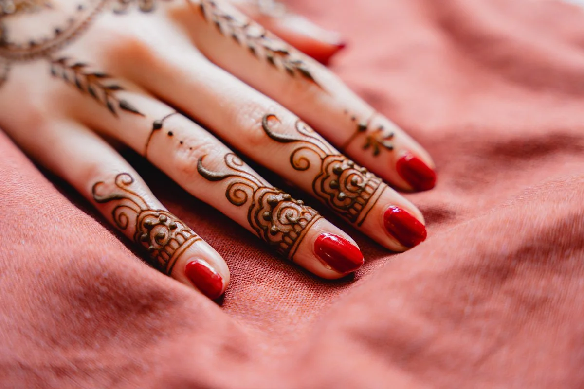 Close-up of a hand with intricate henna tattoos on fingers, bright red nail polish, resting on soft pink fabric.