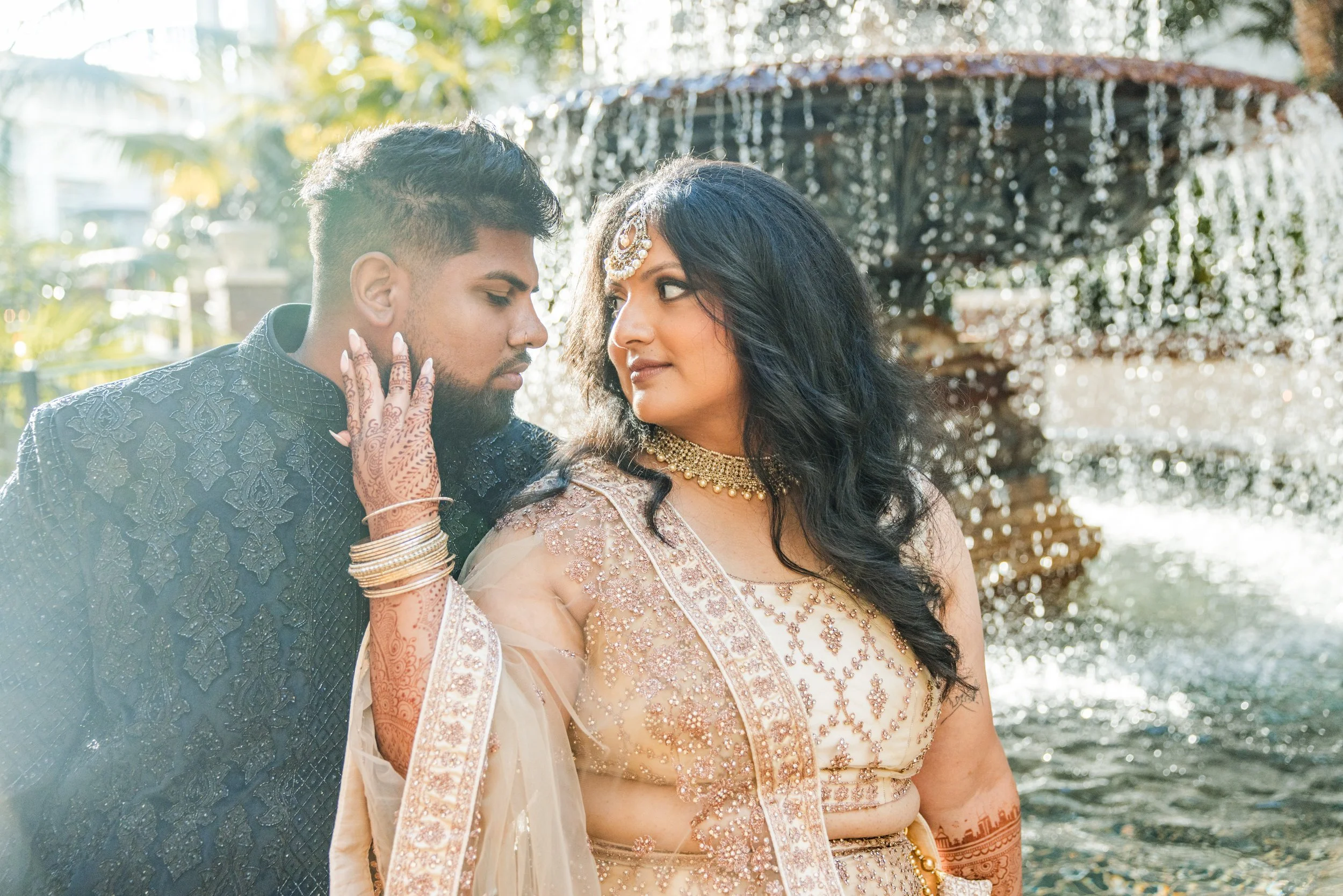 A couple dressed in traditional South Asian wedding attire outdoors, with the groom in a dark embroidered sherwani and the bride in a beige embellished saree, touching foreheads near a water fountain backdrop.