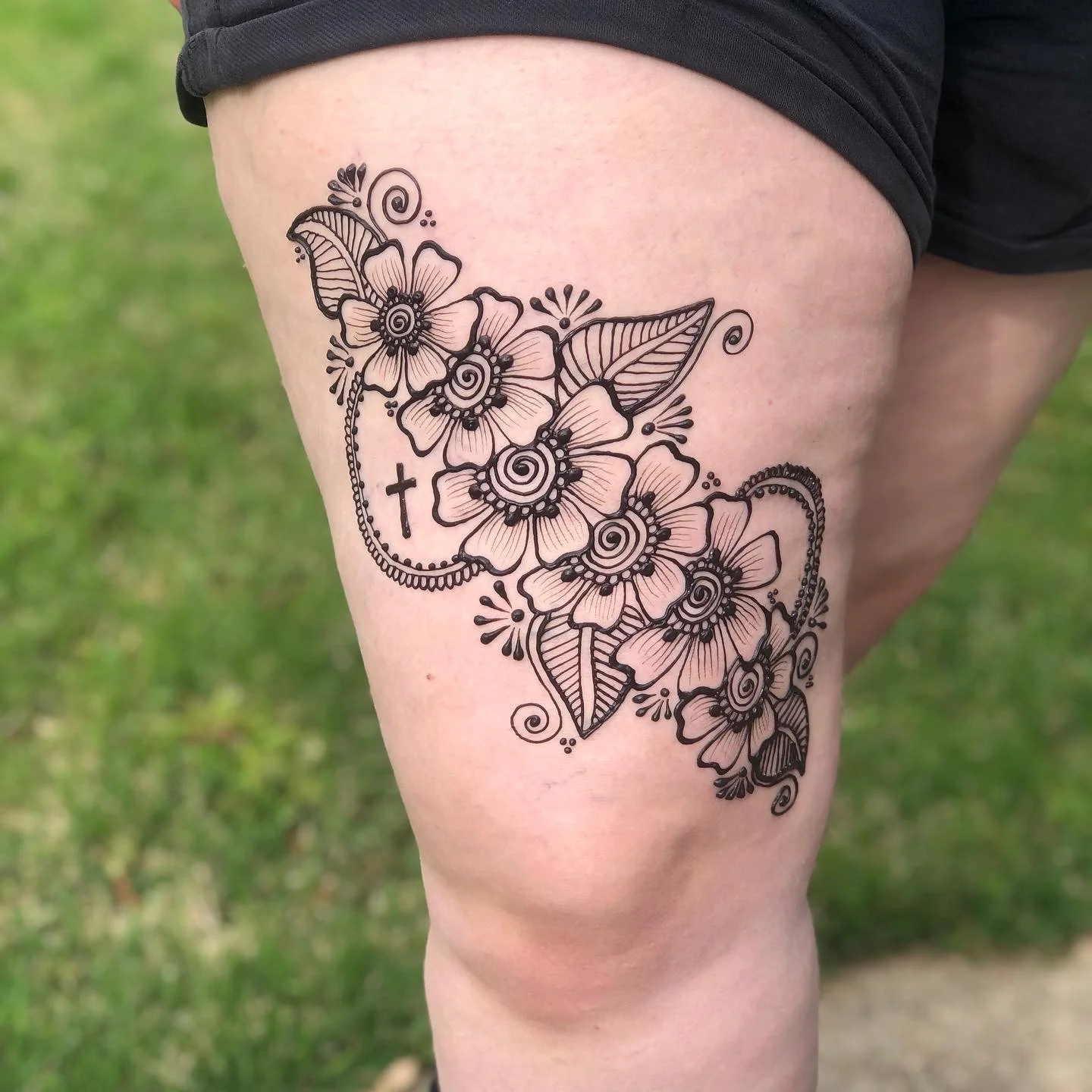 A thigh henna tattoo featuring a floral design with detailed line work, including a cross and decorative elements.
