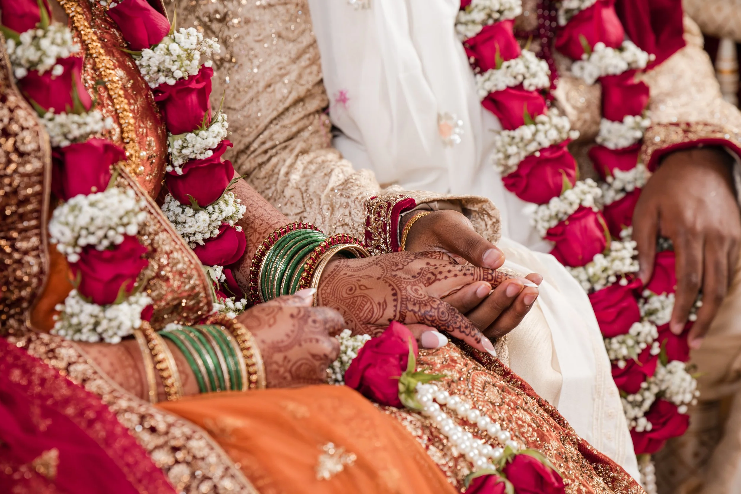 Close-up of a group of women in traditional Indian wedding attire, adorned with colorful jewelry, floral garlands, and intricate henna designs on their hands, sitting together in celebration.