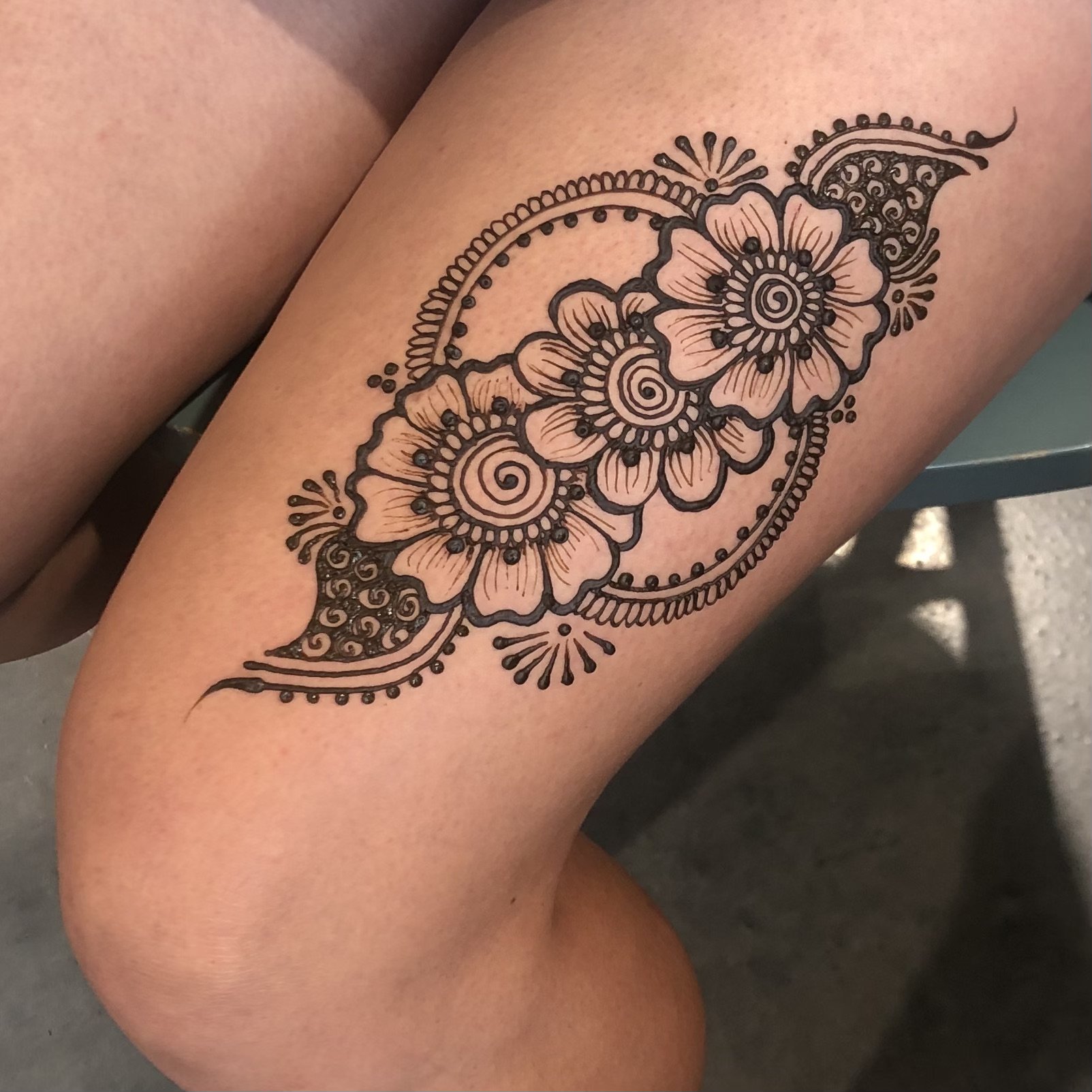 A floral henna tattoo on a person's lower thigh, featuring large flowers, swirling patterns, and decorative accents in black ink.