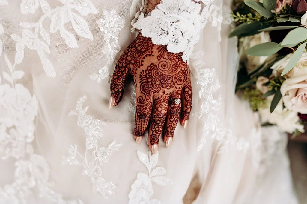Close-up of a woman's hand on her wedding dress, featuring intricate henna designs and a diamond ring, surrounded by delicate lace and floral embellishments.