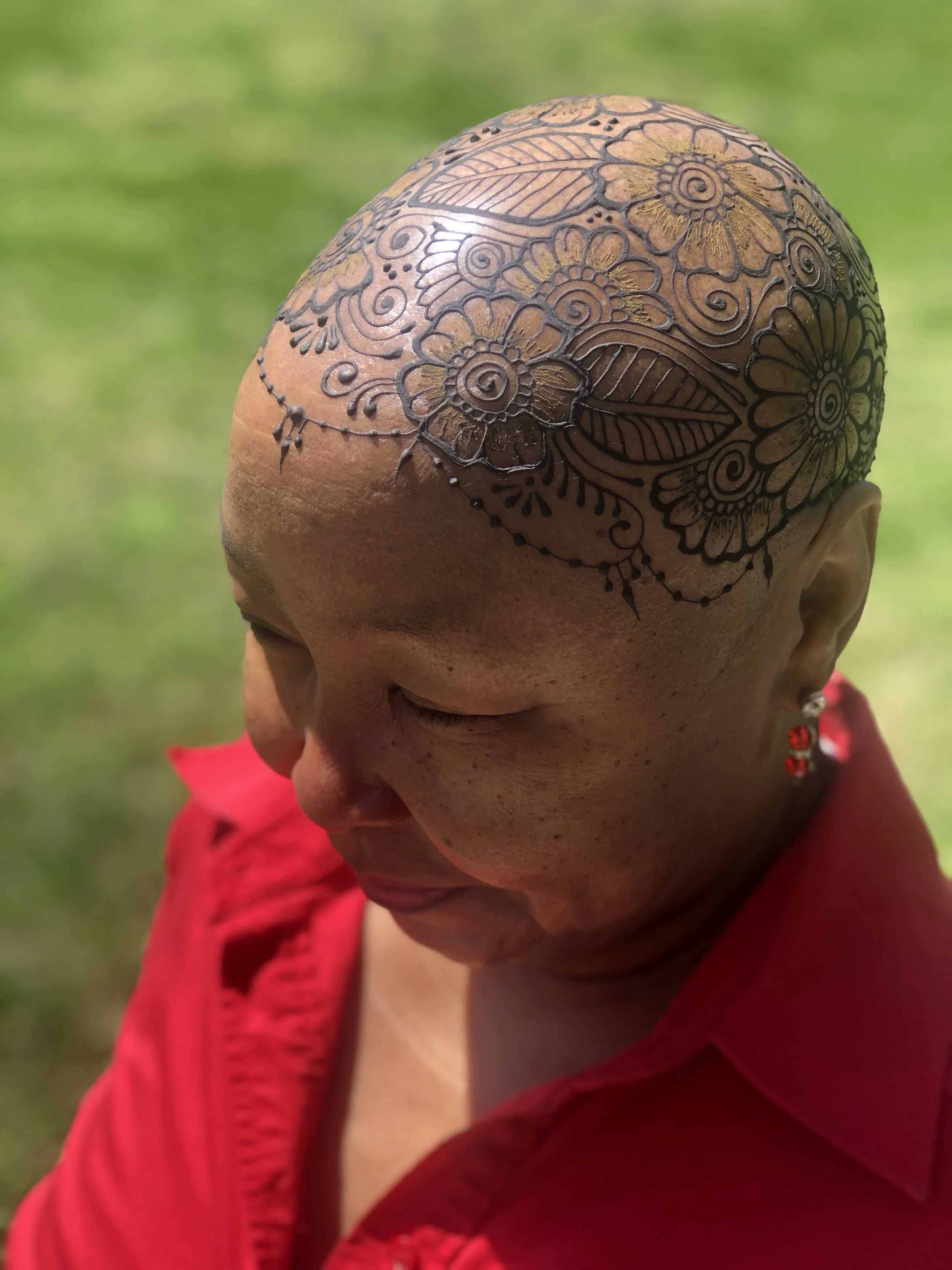 A person with a shaved head decorated with intricate henna or tattoo art featuring flowers, leaves, and swirls, wearing a red garment, outdoors on a grassy area.