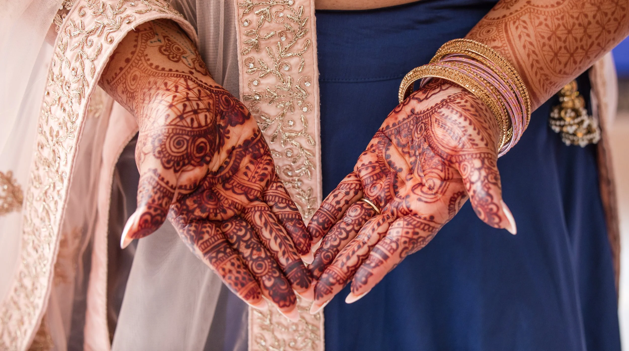 Close-up of a person wearing henna designs on their hands, gold bangles, and traditional Indian clothing with embroidery, standing next to another person in a blue outfit.