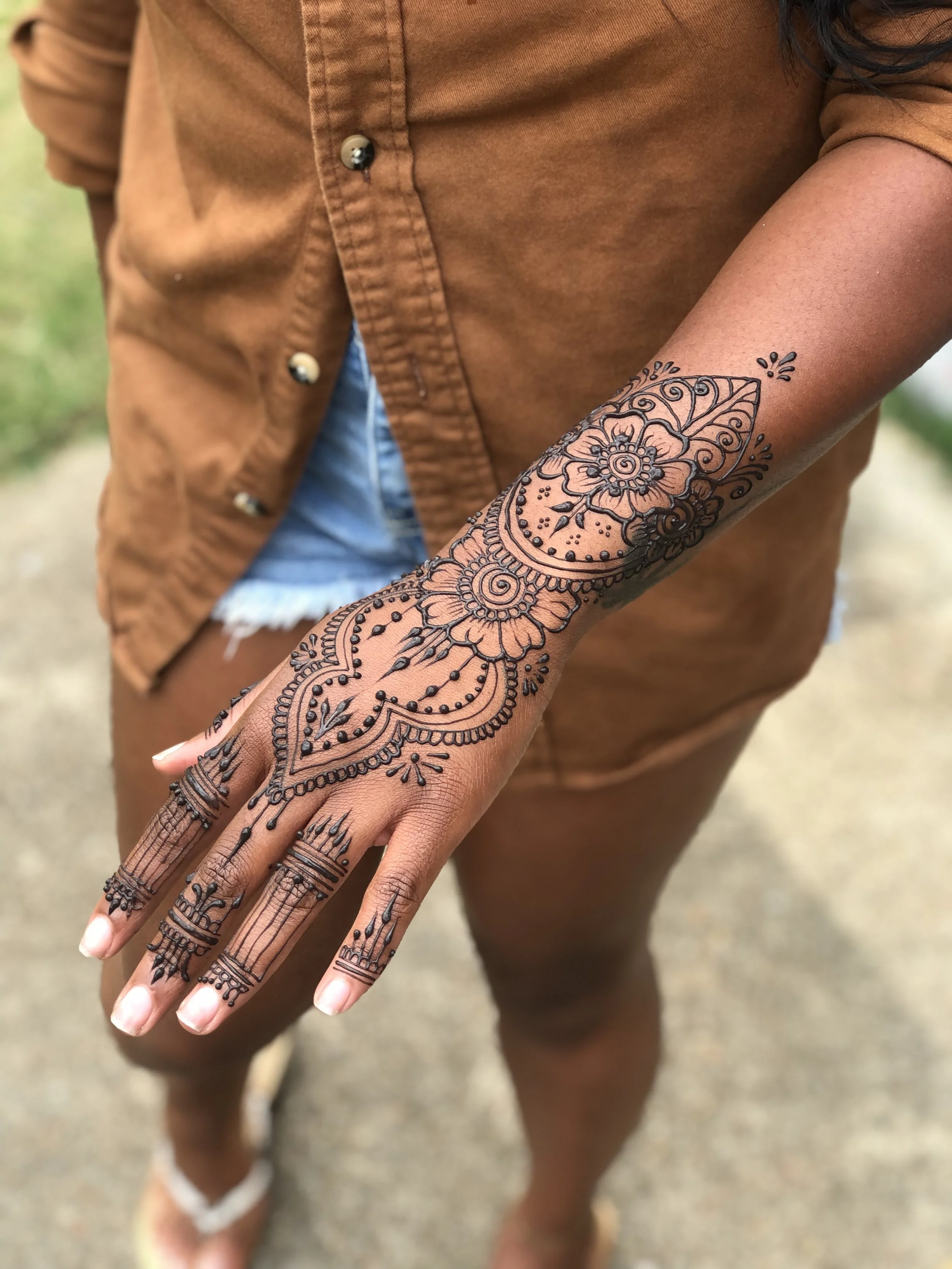 Close-up of a person's arm and hand displaying intricate henna tattoo, with a brown shirt and denim shorts visible in the background.