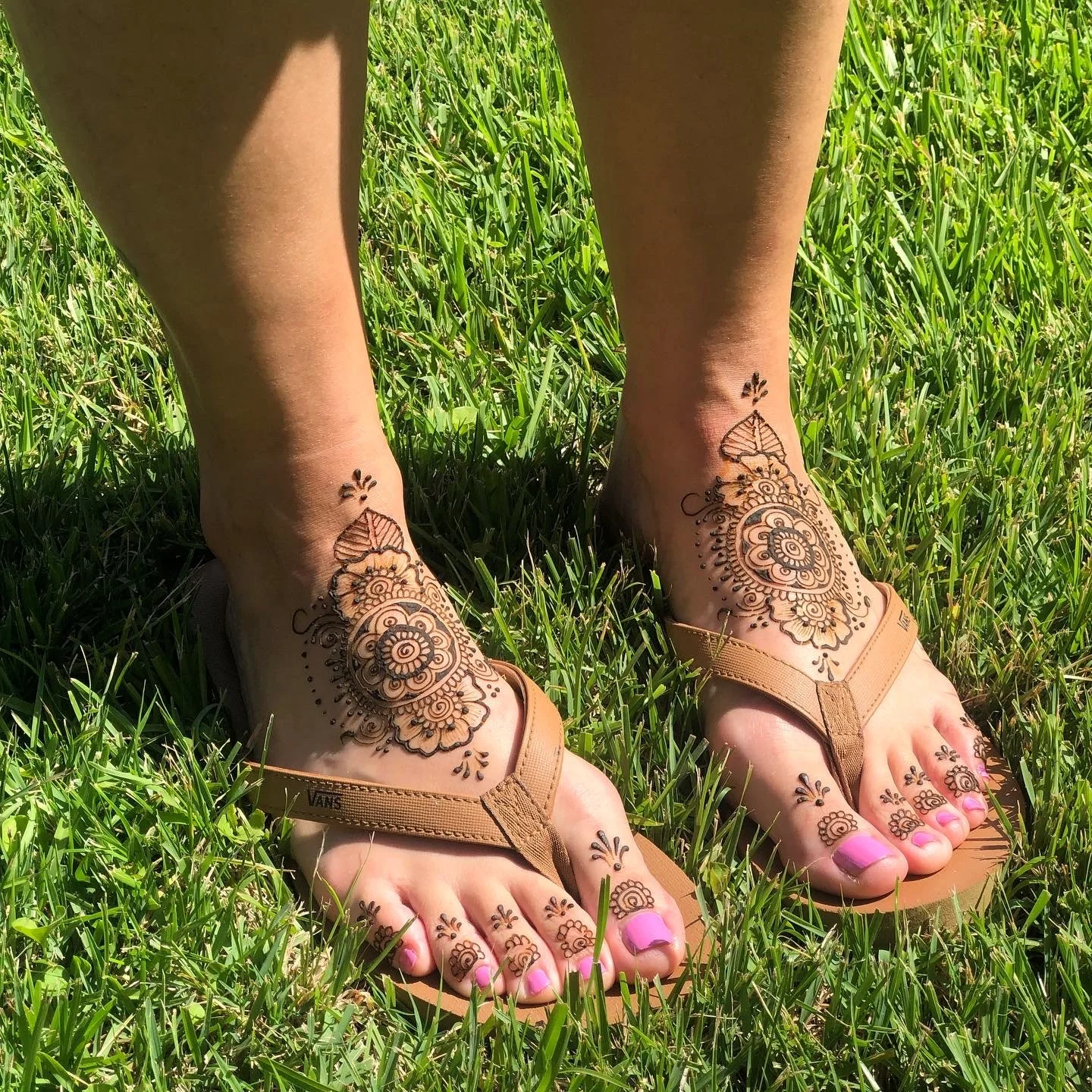 A person's feet in flip-flops on grass, decorated with intricate henna tattoos on the feet and ankles.