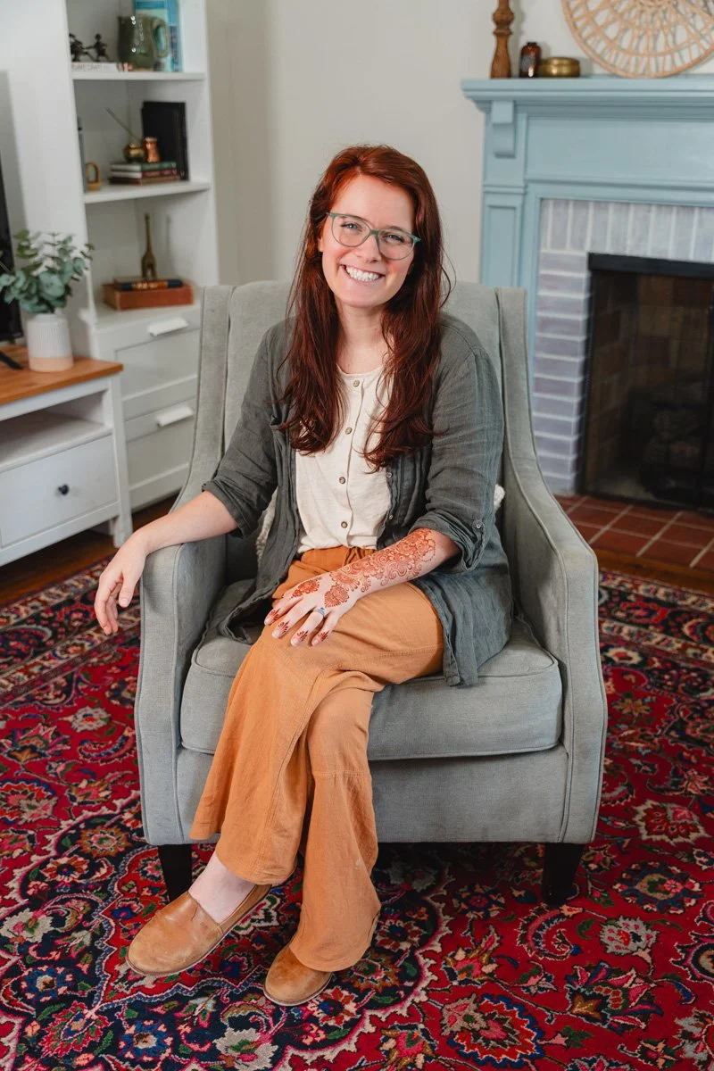 A woman with red hair and glasses sitting in a grey armchair in a living room. She is smiling, wearing a light-colored shirt, tan pants, and a gray cardigan. The room has a patterned rug, a fireplace with a blue mantel, and bookshelves