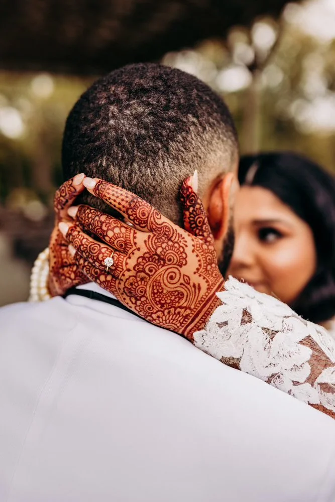 Close-up of a woman with mehndi designs touching the back of a man’s head during a wedding or celebration, with a woman in the background.