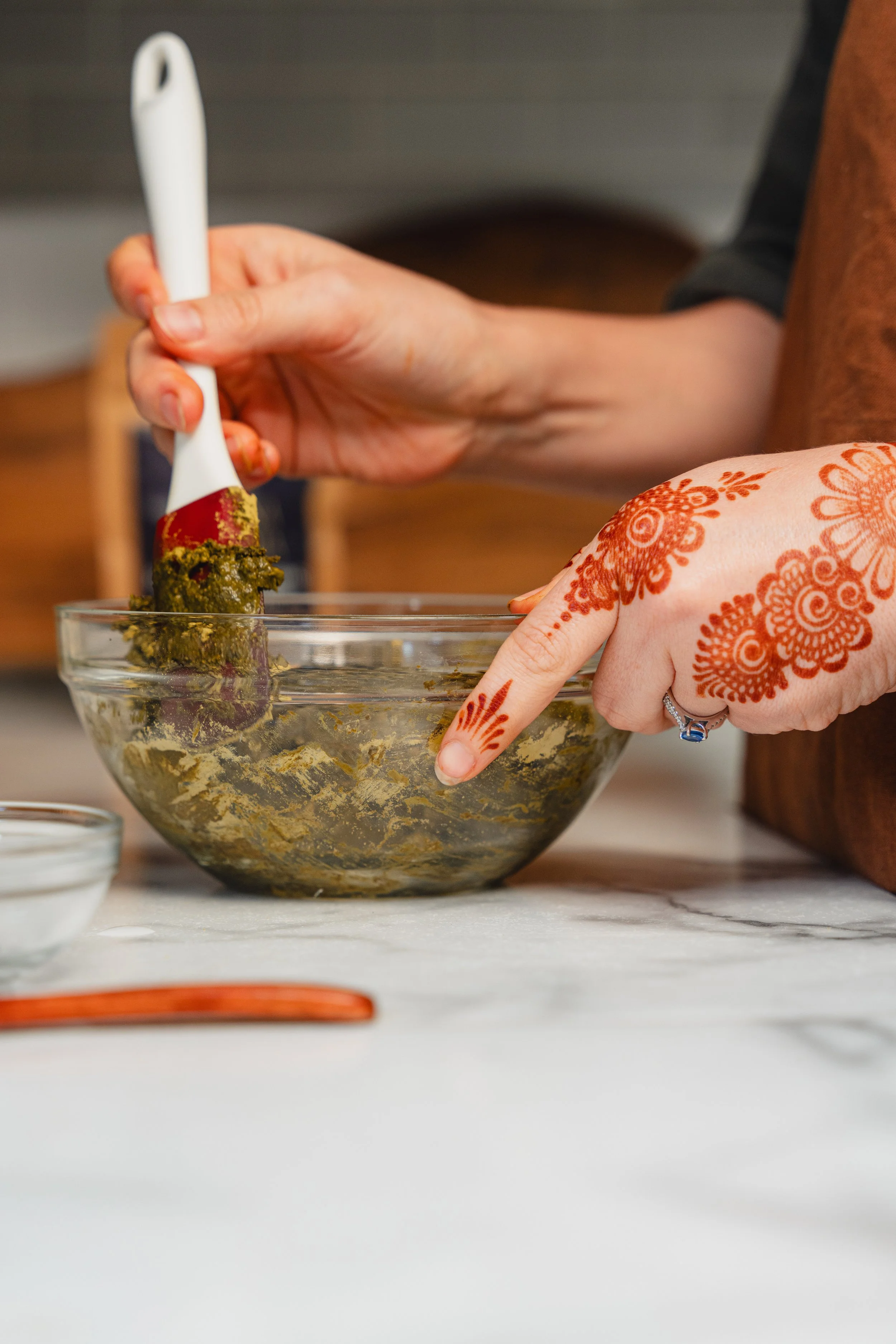 Person with henna decorated hand stirring a bowl of green paste with a white spatula in a kitchen setting.