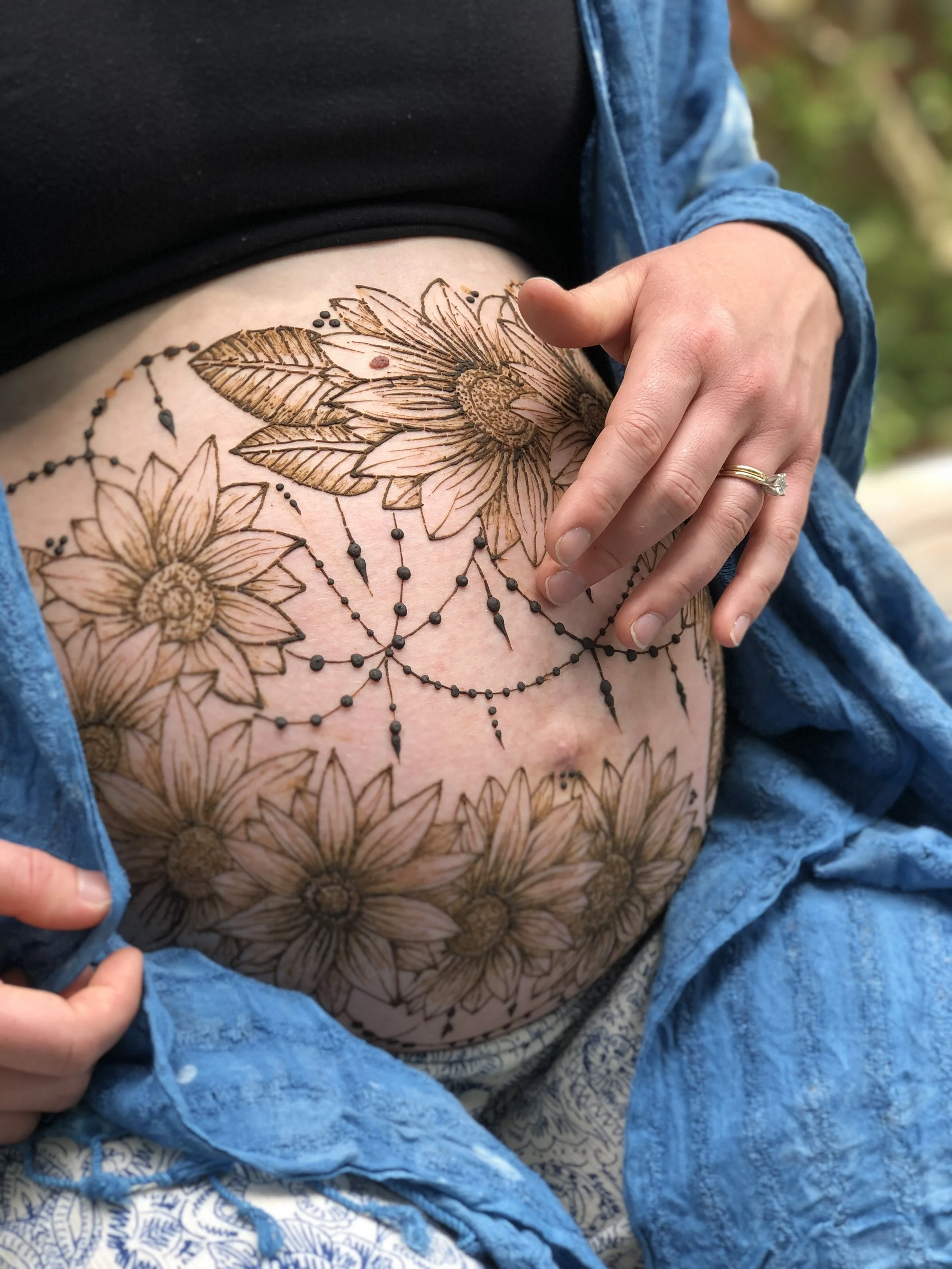 Close-up of a person's torso with a large floral tattoo, partially covered by a blue jacket, and hand with a wedding ring resting on the tattoo.