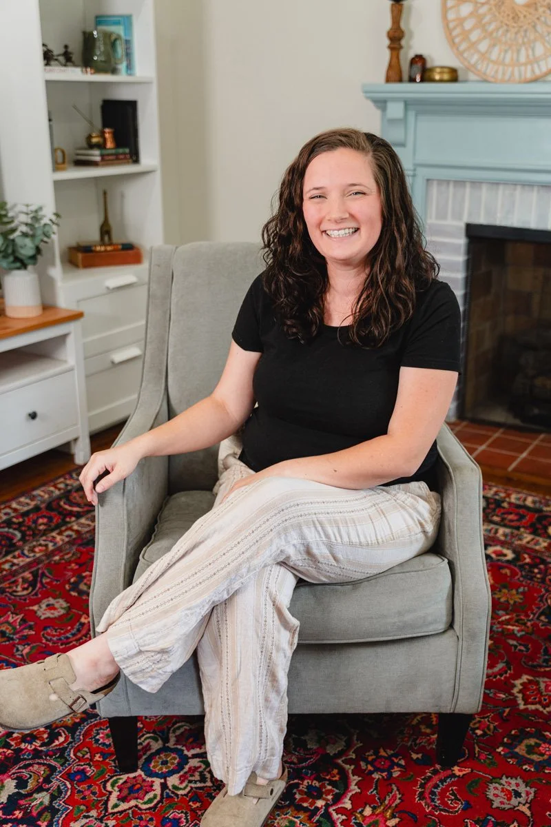 A young woman with curly brown hair sitting on a gray armchair in a living room, smiling at the camera. She is wearing a black t-shirt and beige striped pants, with beige slippers. The background includes a white bookshelf with decorative items, a white side table, and a fireplace with teal mantle and decorative objects on top.