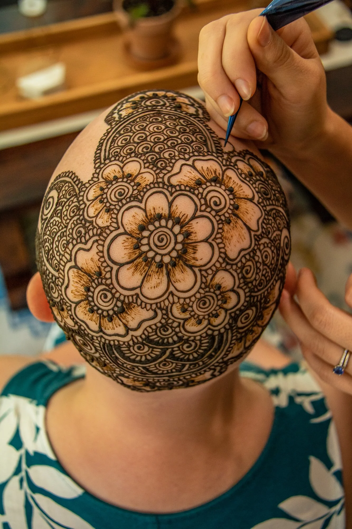Child with a henna tattoo on her scalp, featuring floral and swirl patterns, while a person applies the henna design with a fine-tipped applicator.