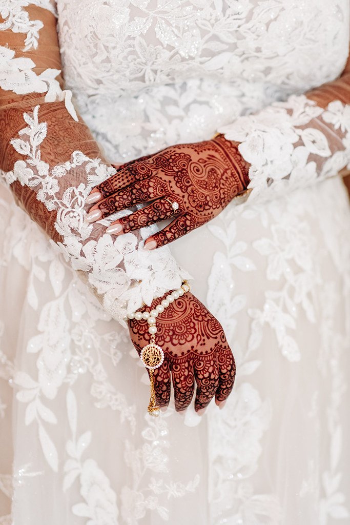 Close-up of a bride's hands with detailed mehndi (henna) designs, wearing a pearl bracelet and ring, dressed in a white lace wedding gown.