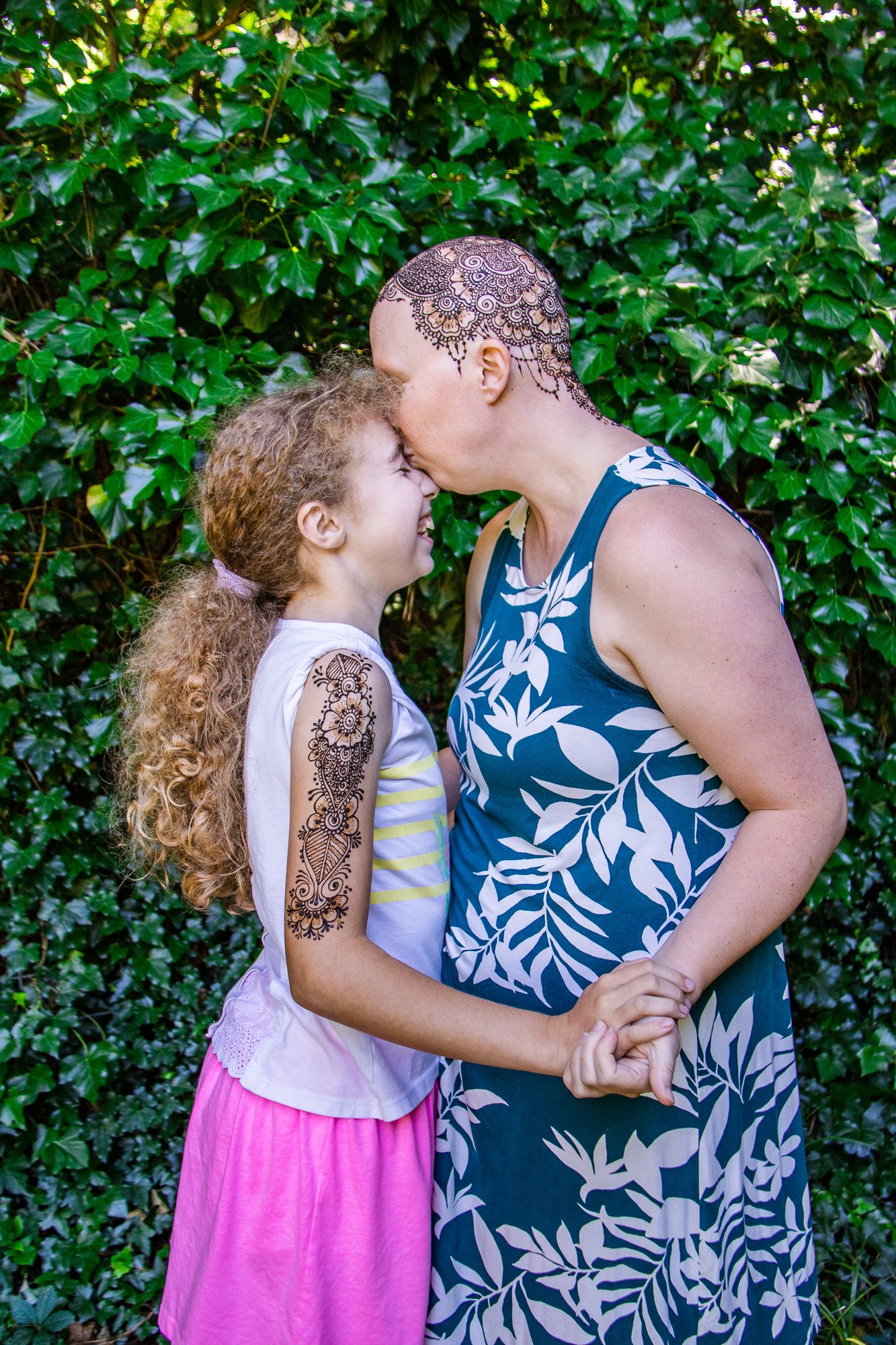 A woman and a girl with henna tattoos on their arms and head sharing a kiss on the forehead while holding hands in front of green leafy bushes.