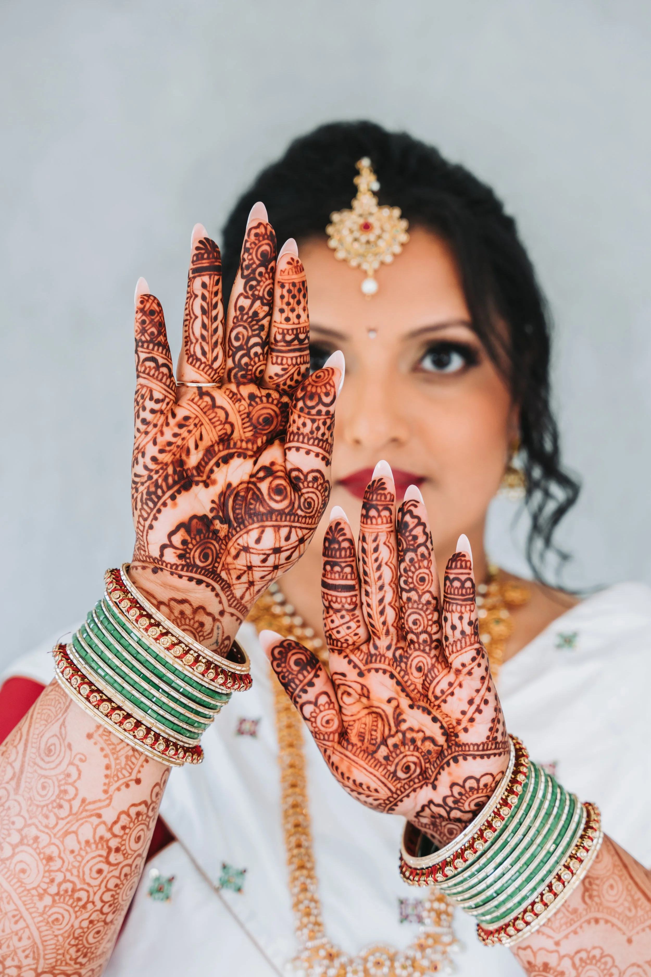 Close-up of a woman with henna on her hands and traditional Indian jewelry, raising her hands with detailed henna designs, wearing colorful bangles and a white saree with gold embroidery.