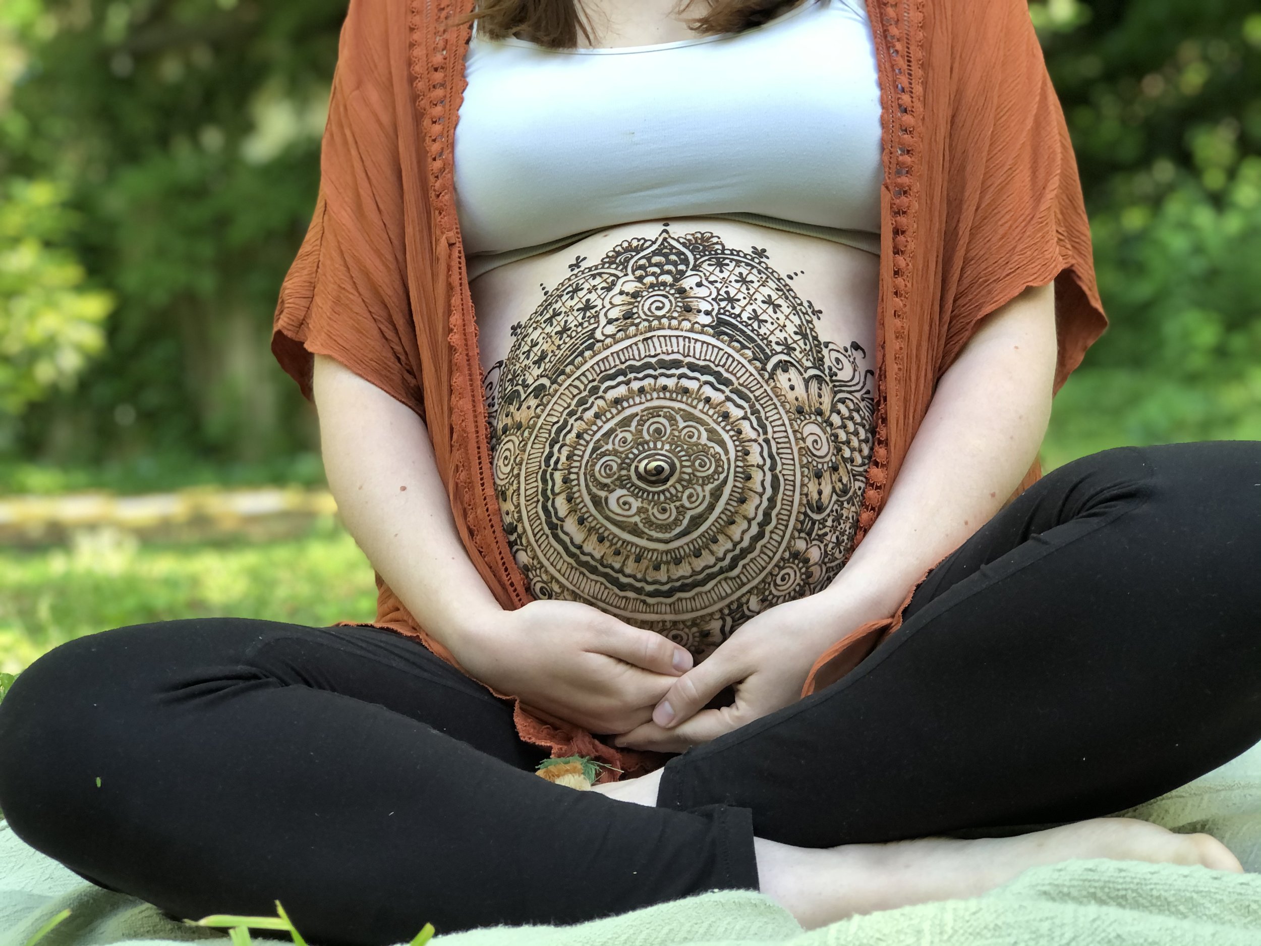 Close-up of a woman's midsection showing a large, intricate black and gold henna tattoo on her stomach. She is wearing a white top and an orange lace shawl, sitting outdoors on a green surface with a blurred background of trees and grass.