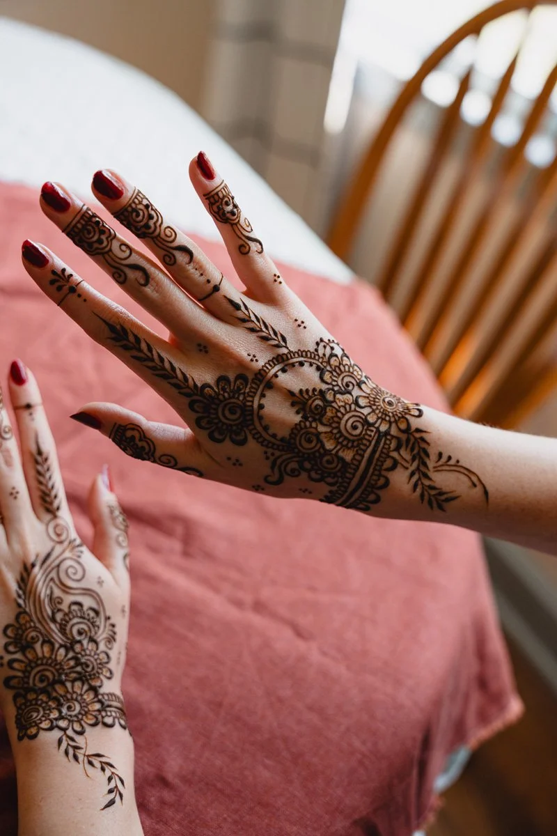 Close-up of hands with intricate henna designs, red painted nails, resting on a pink fabric surface, with a wooden chair in the background.
