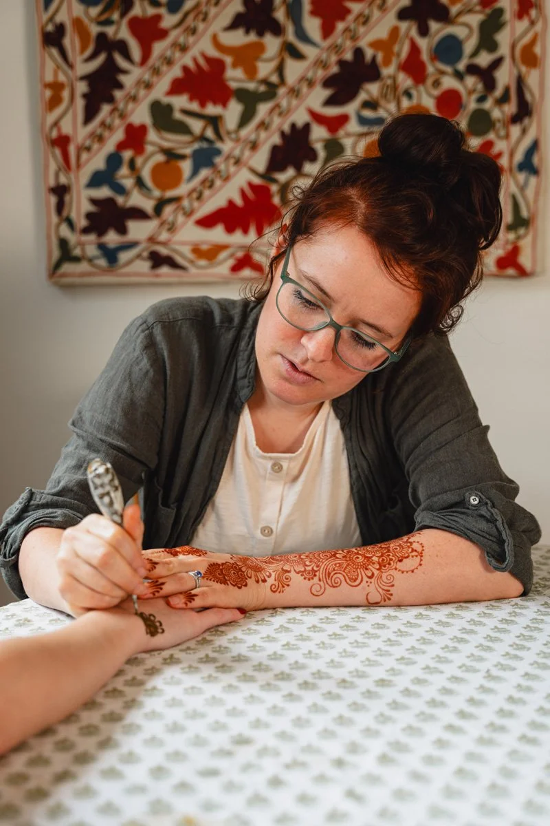 A woman with glasses and dark hair styled in a bun is decorating a person's hand with mehndi or henna at a table covered with a patterned cloth. A colorful, intricate textile hangs on the wall behind her.