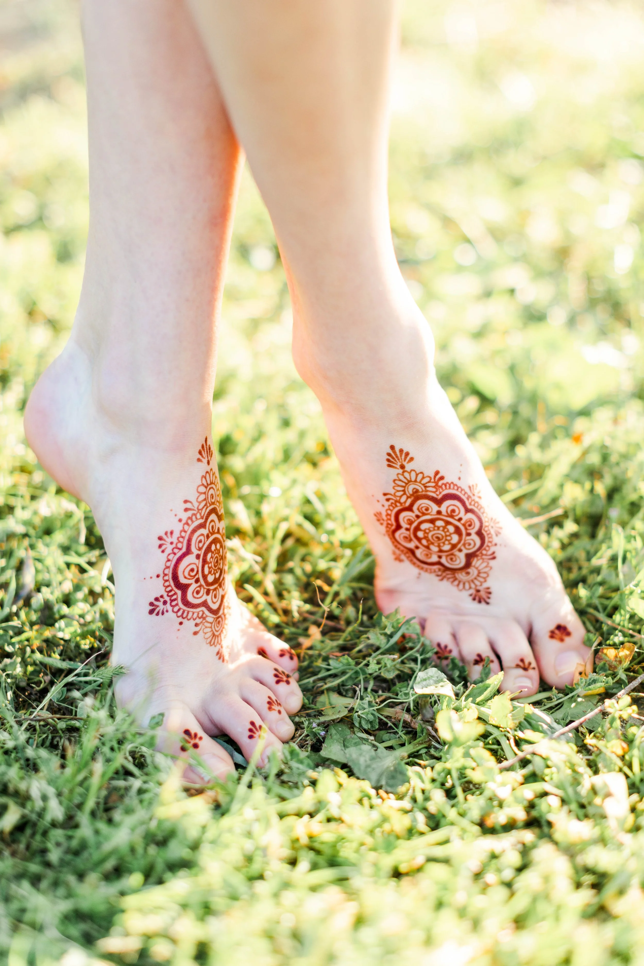 Close-up of bare feet with henna designs standing on grass and greenery outdoors.