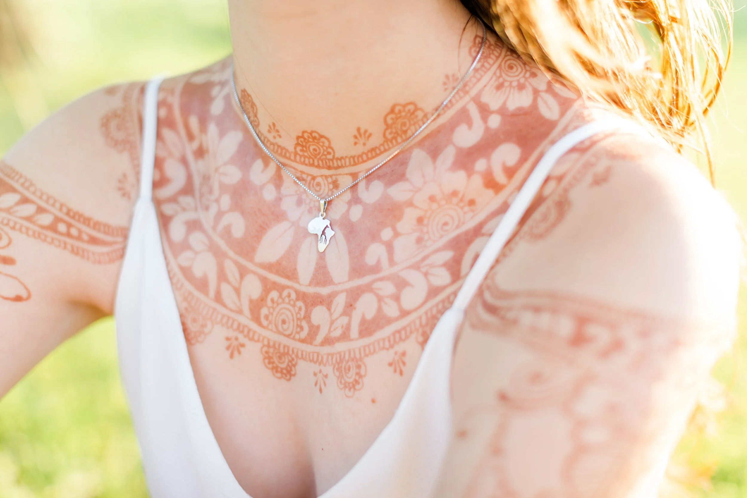 Close-up of a woman's shoulder and neck showcasing detailed orange henna tattoos, with a silver necklace featuring a skull pendant, against a blurred outdoor background.