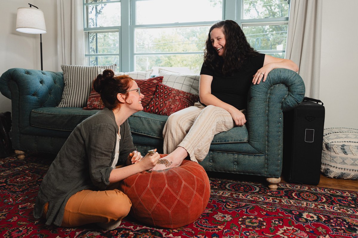 Two women, one sitting on a teal couch and the other on a red bean bag, smiling and laughing together in a cozy living room with large windows and a patterned rug.