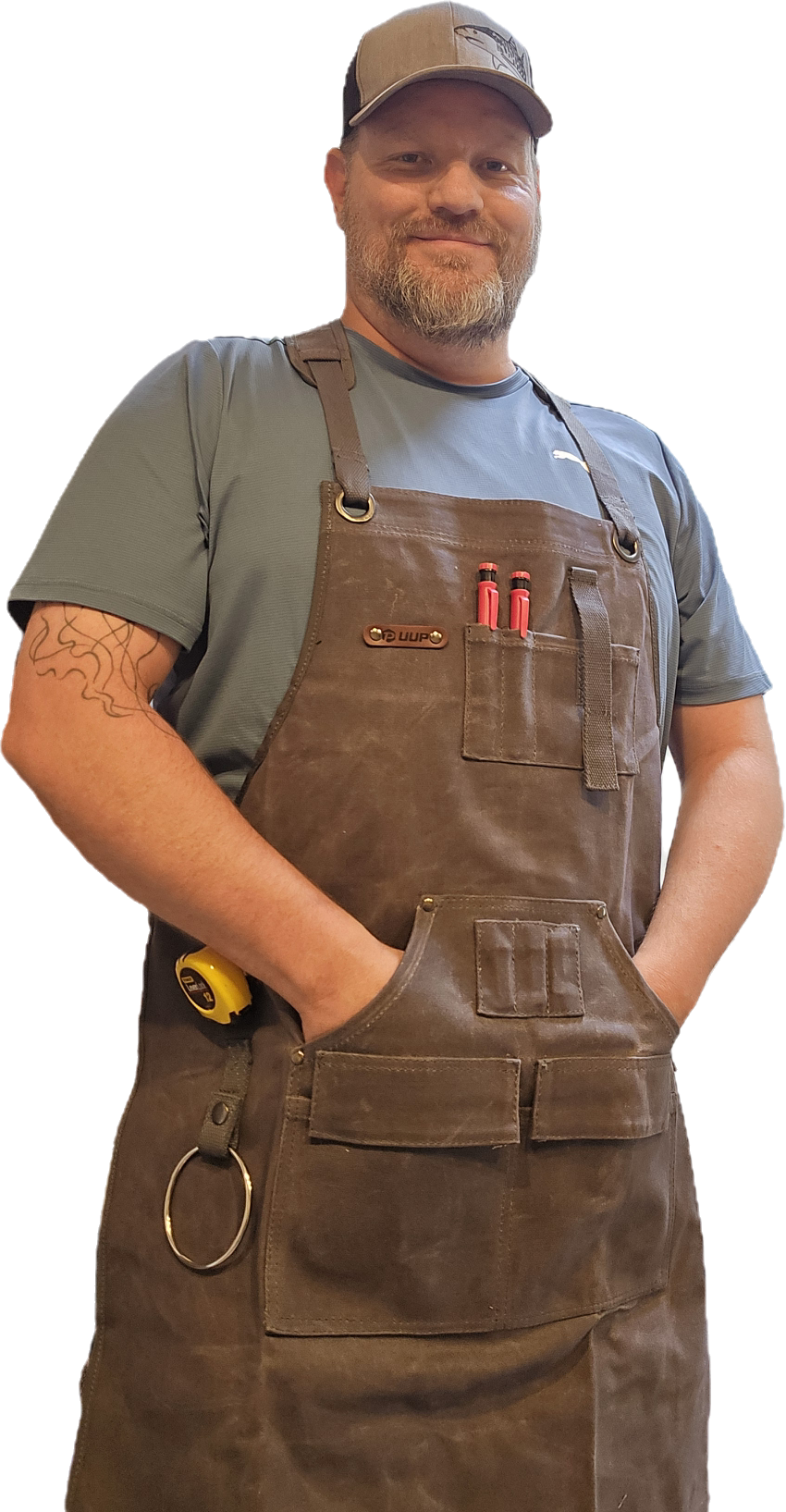 A man with a beard wearing a cap, gray T-shirt, and brown leather apron with tools in the pocket, standing with hands in pockets, smiling.