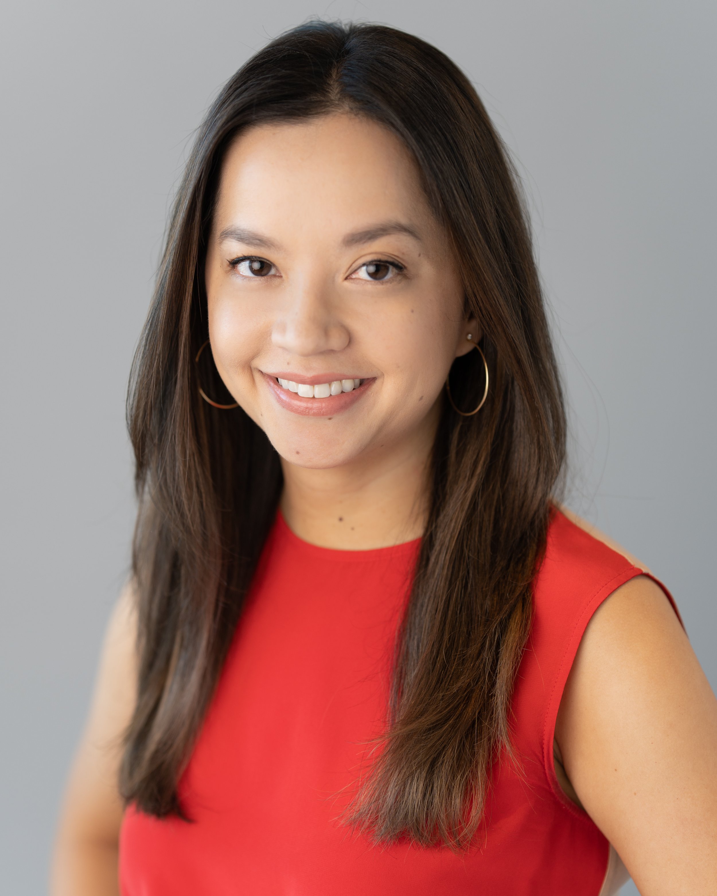 Portrait of a young woman with long dark hair, wearing a red sleeveless top and large gold hoop earrings, smiling softly against a plain gray background.