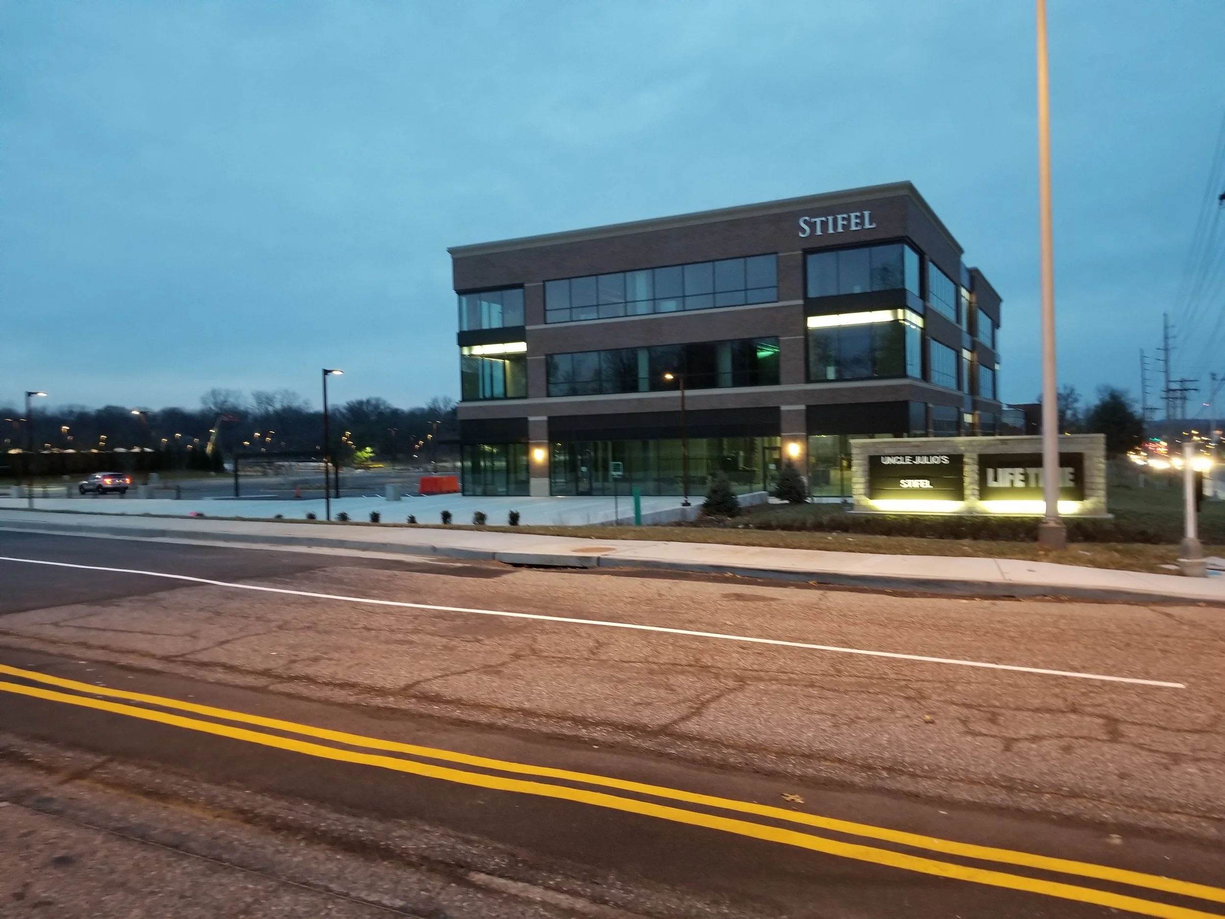 A modern three-story office building with the name 'STIFEL' on the corner. In front, a sign reads 'UNCLE JULIO'S, STIFEL, LIFE TIME.' The scene is at dusk with streetlights on, and the building is lit from within.