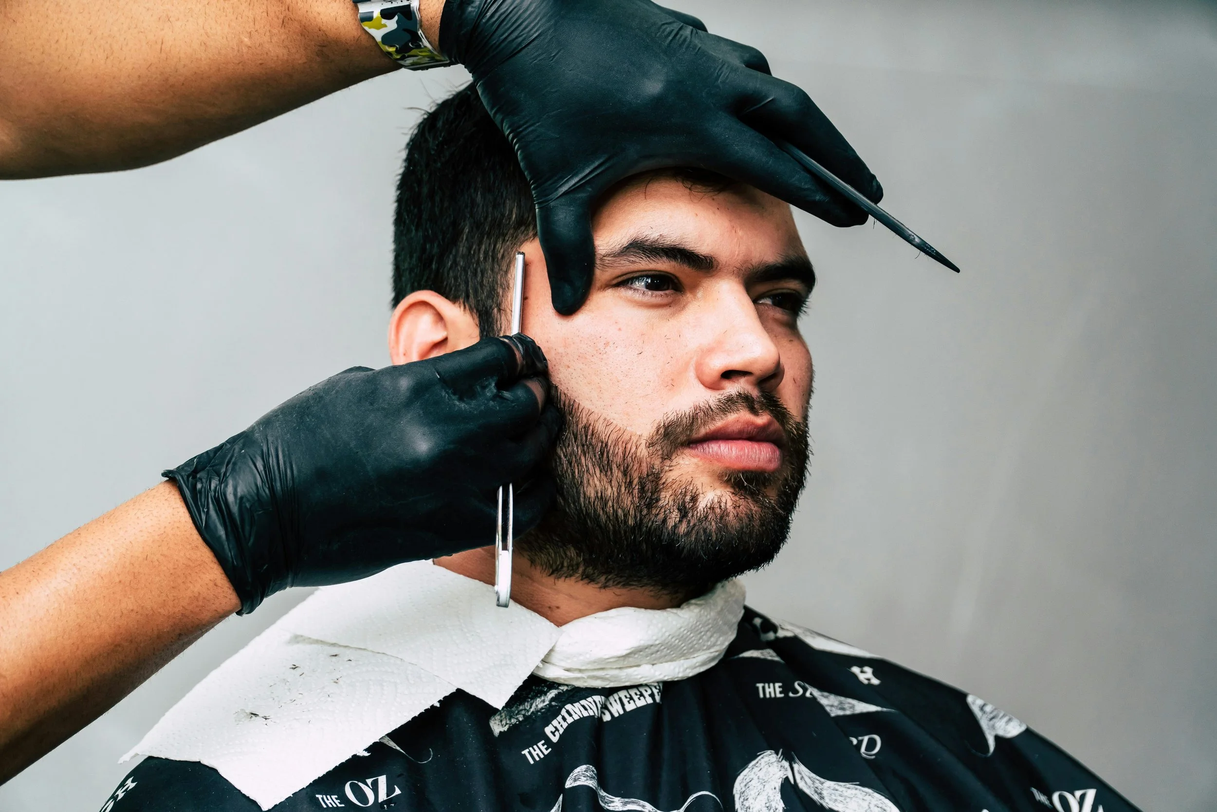 A man with a beard getting a haircut or beard trimming from a barber wearing black gloves, in a barbershop.