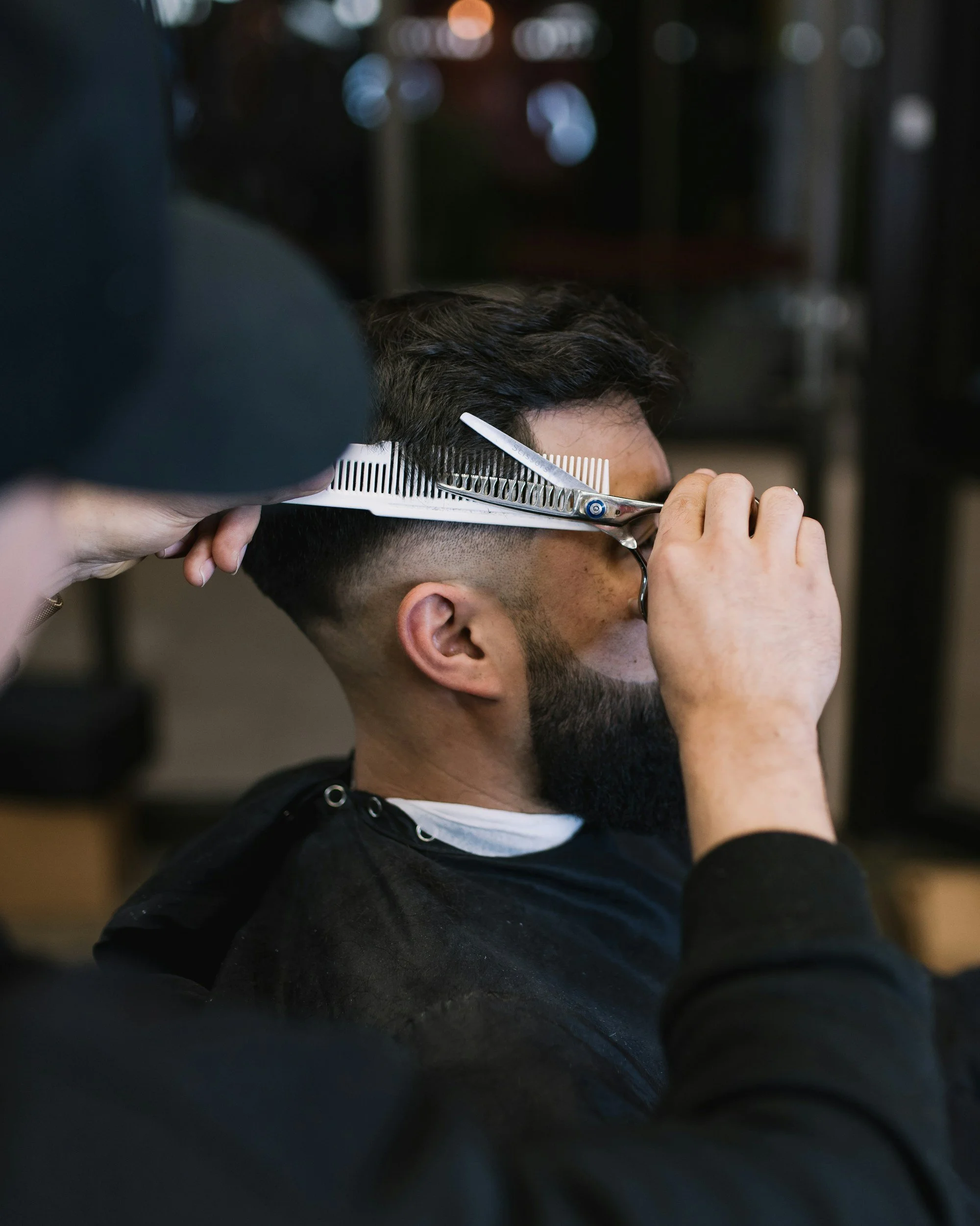 A barber is cutting a man's hair using scissors and a comb, while the man sits with safety glasses on and a black cape over his clothes.