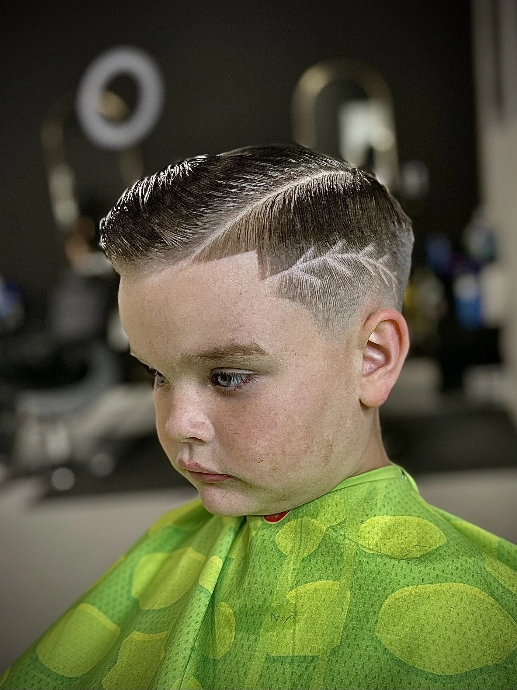 A young boy with a freshly styled haircut at a barbershop, wearing a bright green cape with a leaf pattern.