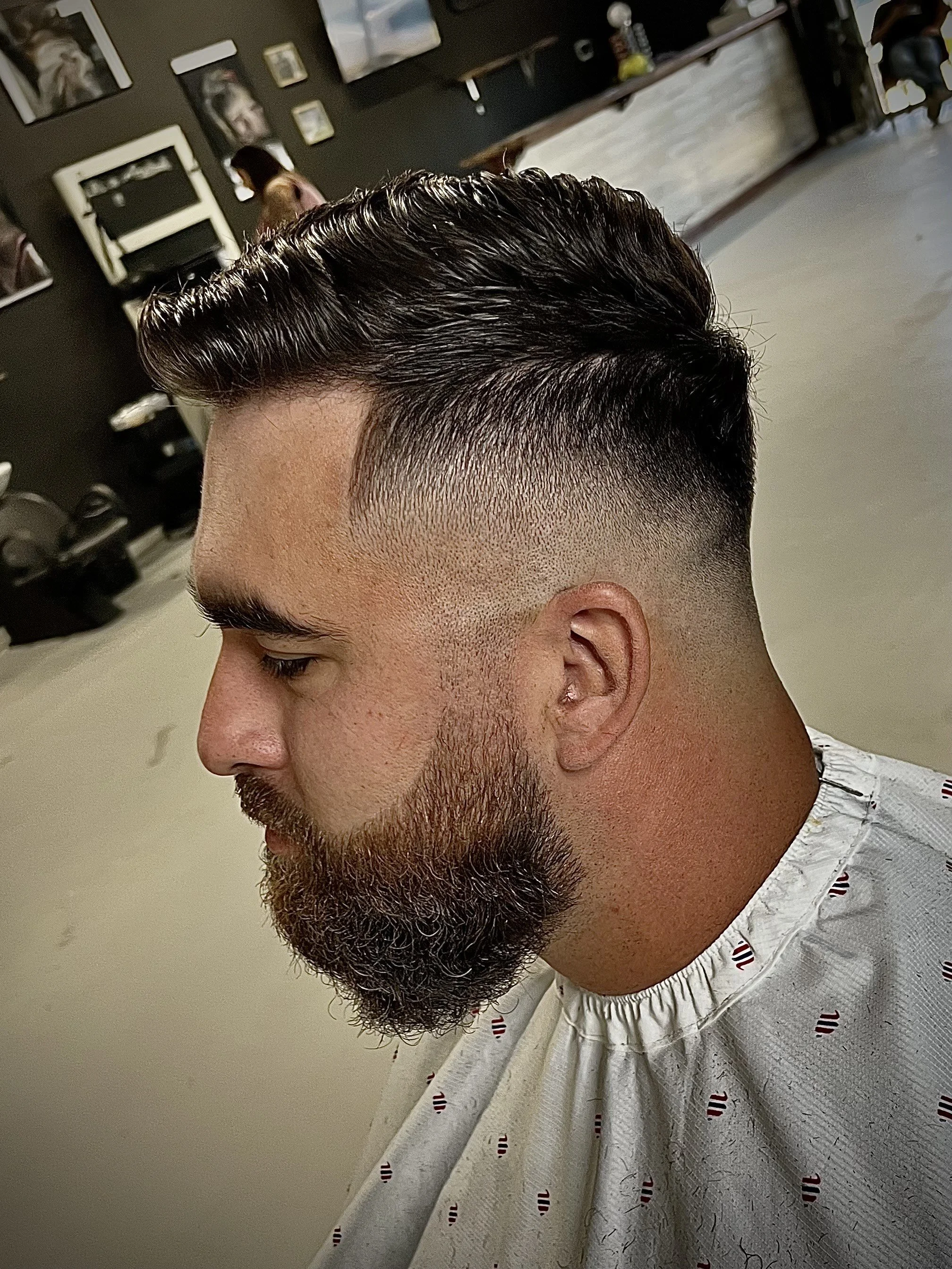 Close-up of a man with a freshly styled haircut featuring a high fade and slicked-back top, with a beard, sitting in a barbershop chair.