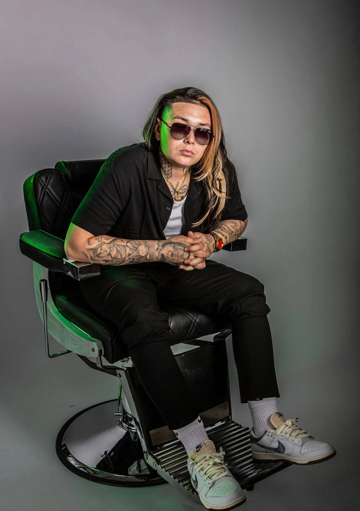 A person with long hair, tattoos on arms, wearing sunglasses, a black shirt, black pants, and sneakers, sitting on a black leather chair in front of a plain grey background.