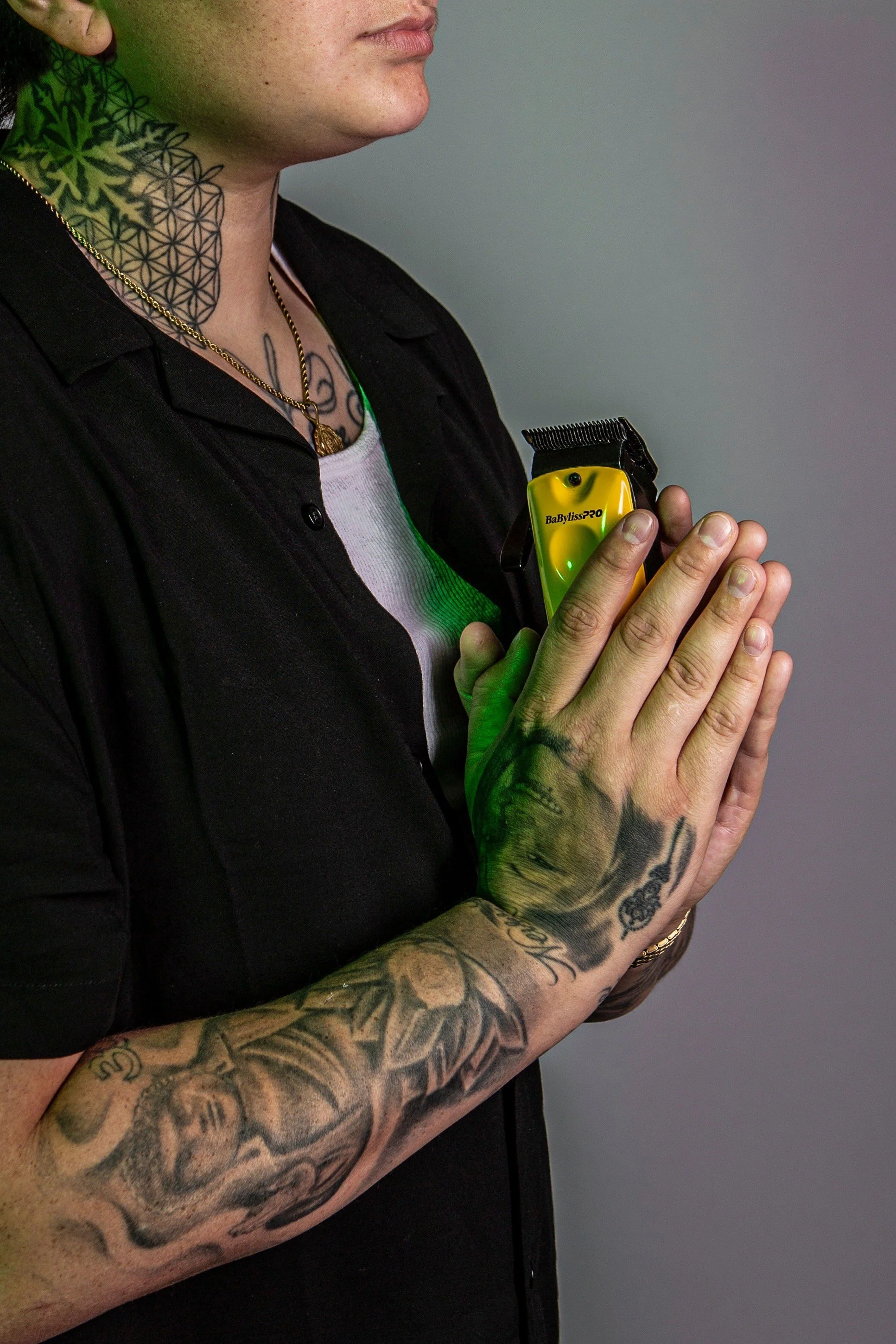 A person with tattoos on their arm and neck holding a yellow and black electric hair trimmer in their hands, pressed together in a prayer-like position.