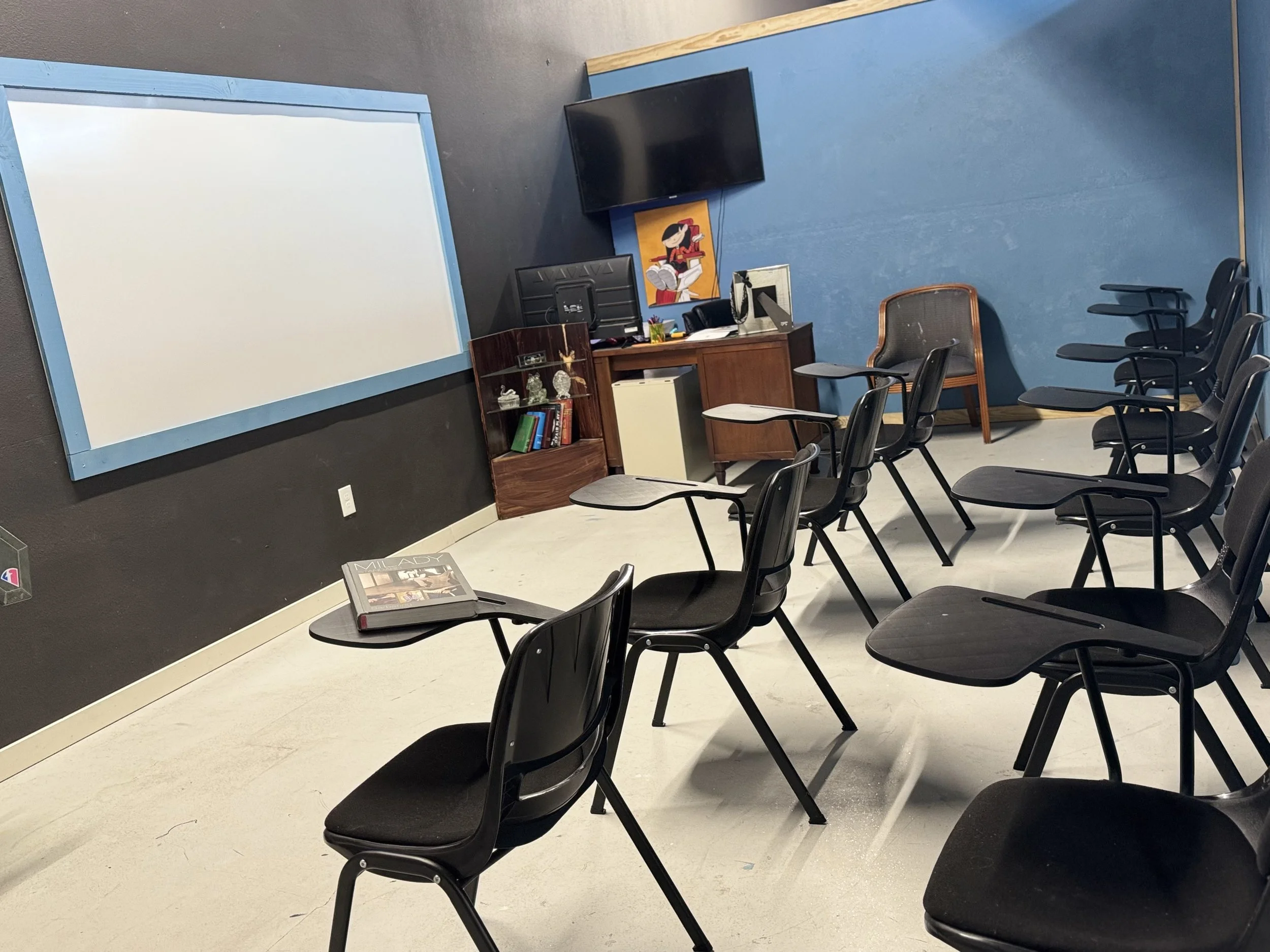 Empty classroom or meeting room with black chairs with attached writing surfaces arranged in rows. A large whiteboard or screen, a desk with a computer and pictures, and a TV mounted on the wall are visible, with a colorful painting nearby.