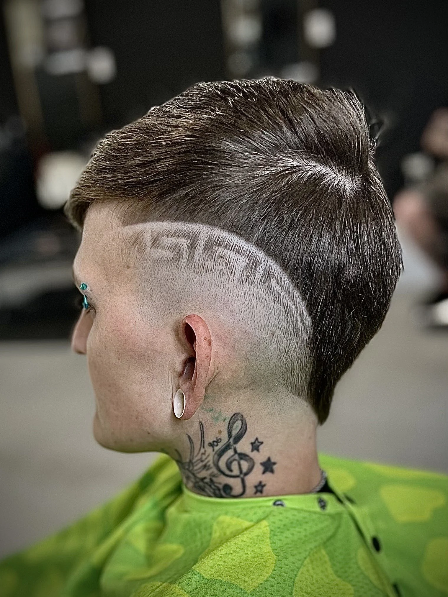 A woman with a short, undercut hairstyle featuring a design shaved into the side. She has tattoos on her neck and is wearing earrings and a bright green shirt.