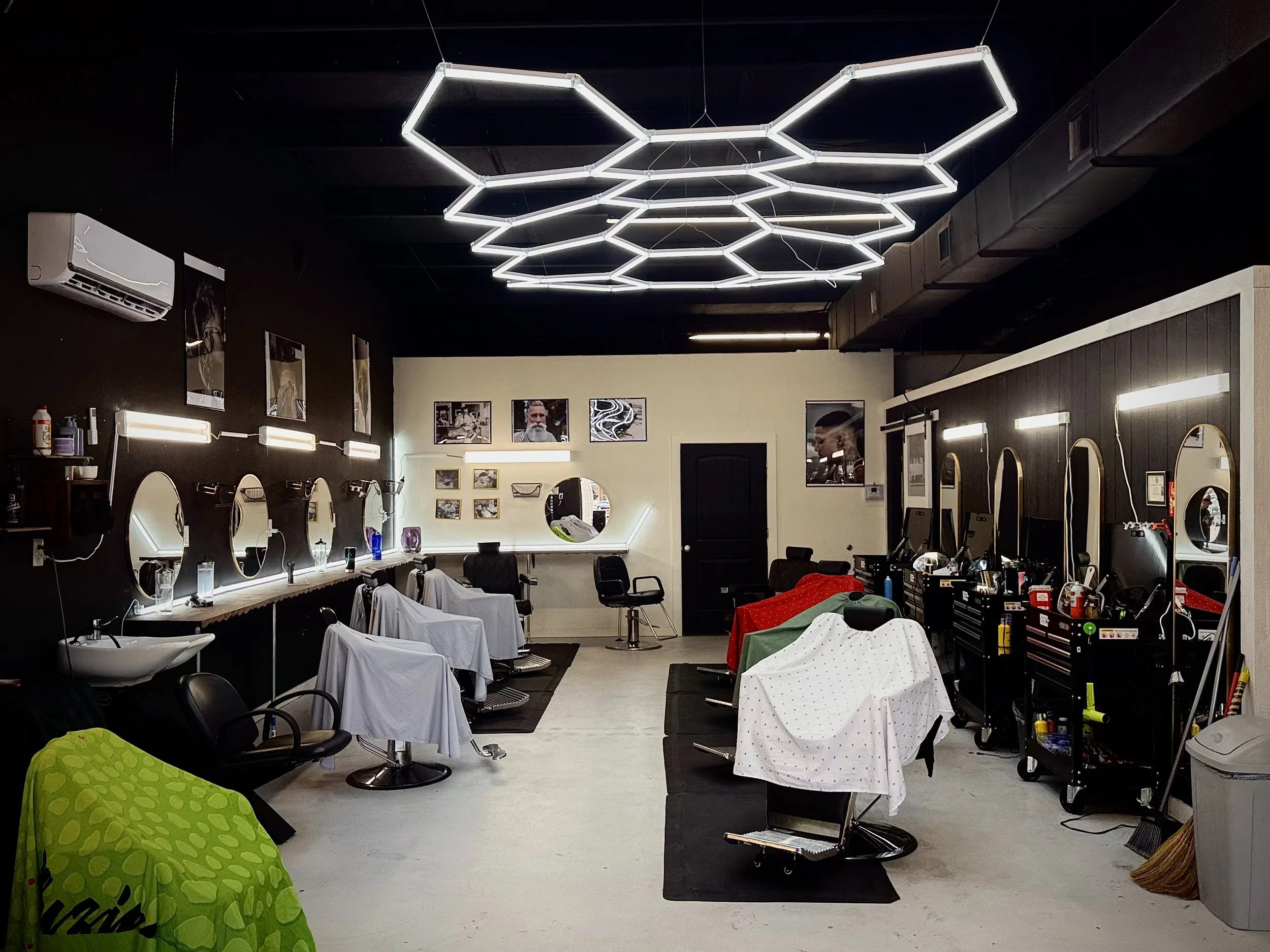 Interior of a modern barbershop with multiple chairs covered in white and black capes, mirrors along the walls, and contemporary hexagonal LED ceiling lights.