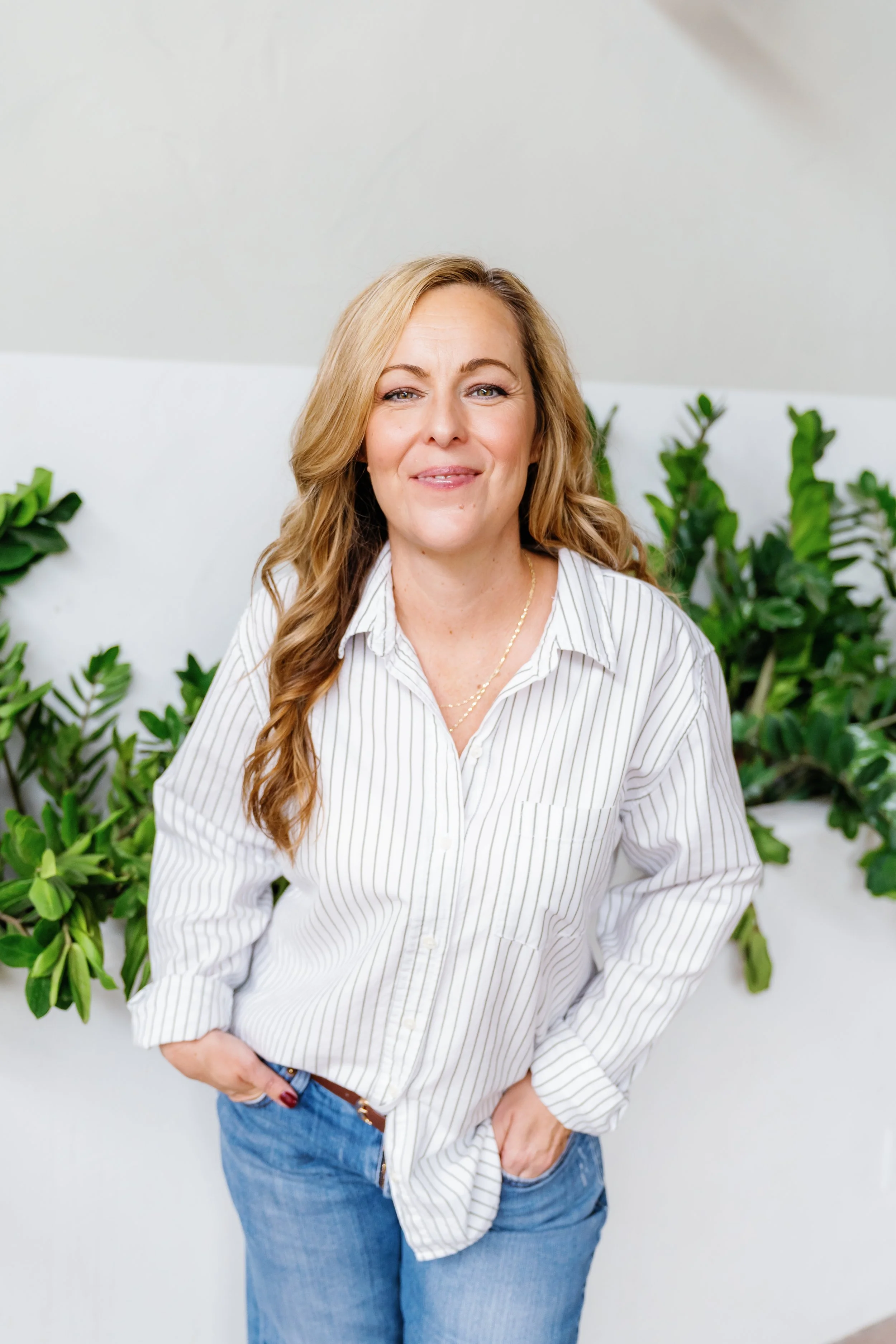 Smiling person in striped shirt and jeans standing in front of green plants.