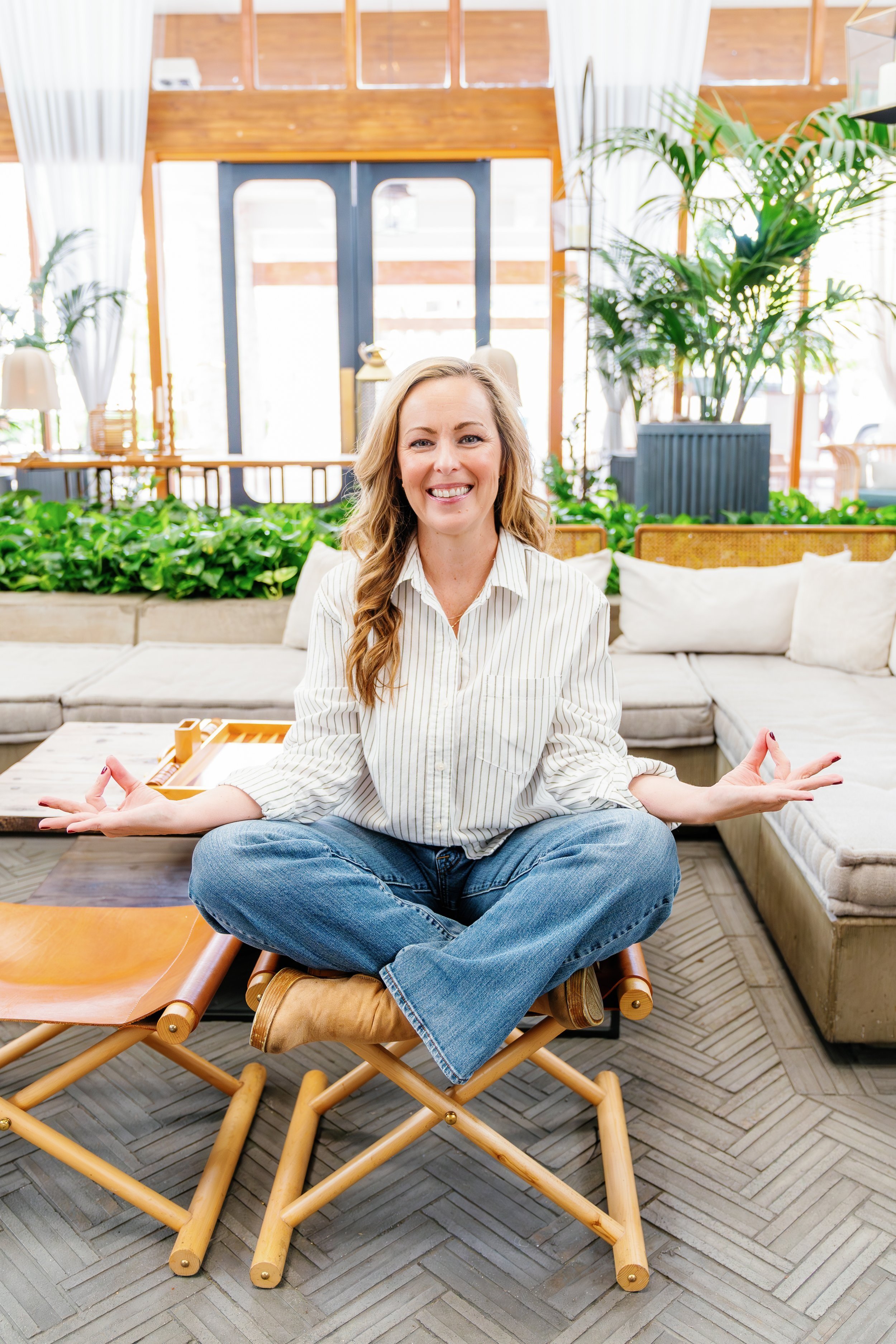 A woman sitting cross-legged on a wooden chair indoors, wearing a striped shirt and jeans, smiling, with houseplants and a cozy seating area in the background.