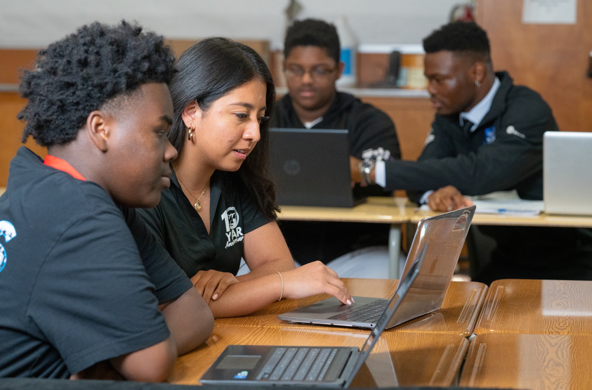 Four students sitting at a wooden table with laptops, focusing on their work in a classroom.