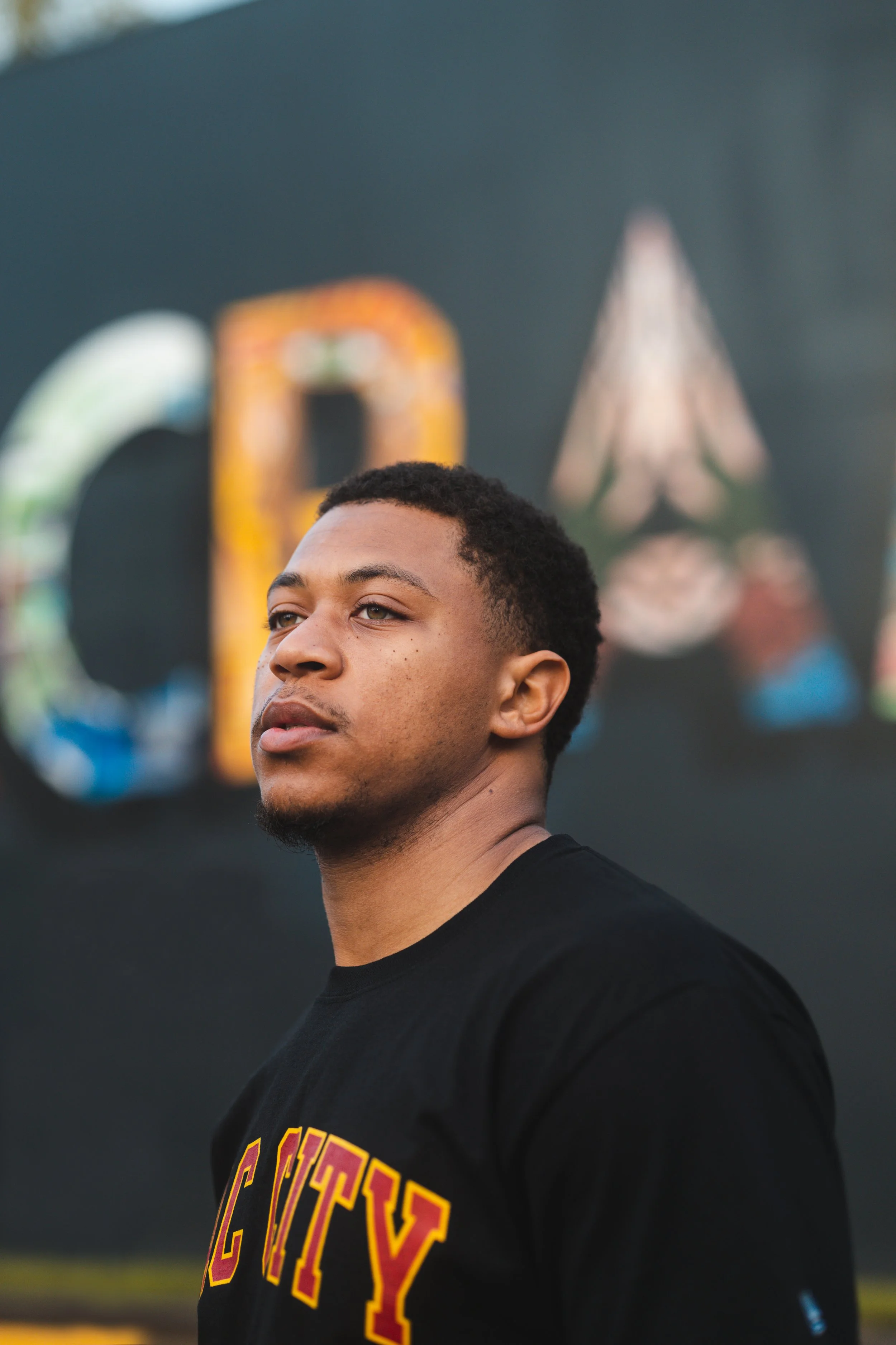 Close-up of a young man with short hair, wearing a black shirt with partially visible yellow and red text, standing outdoors with a blurred background of a rainbow-colored sign or artwork.