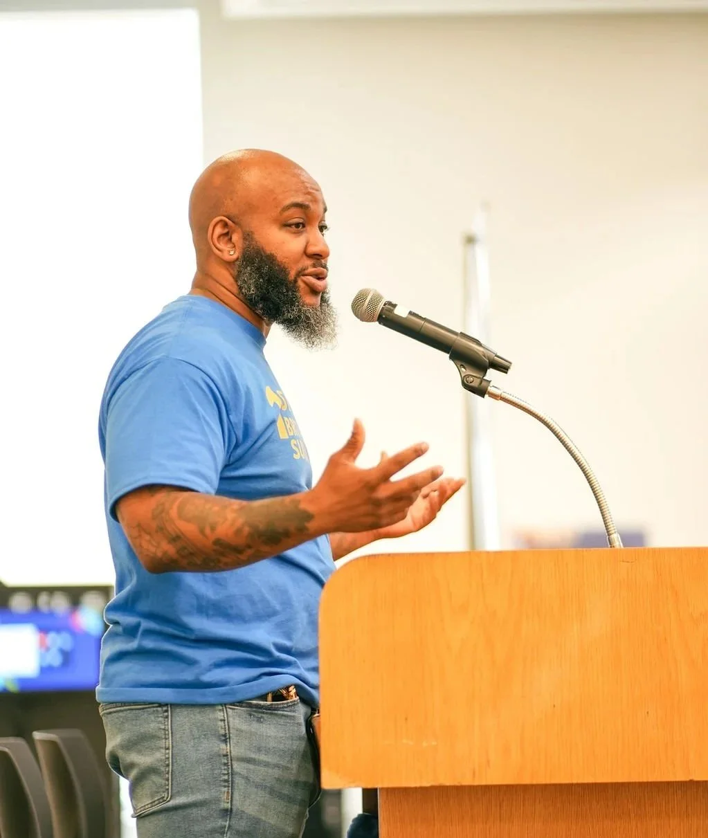 Executive Director of IYT Minnesota addresses the crowd at the first annual Brotherhood Conference in Minnesota. He is wearing a blue T-shirt, standing at a podium and speaking into a microphone.