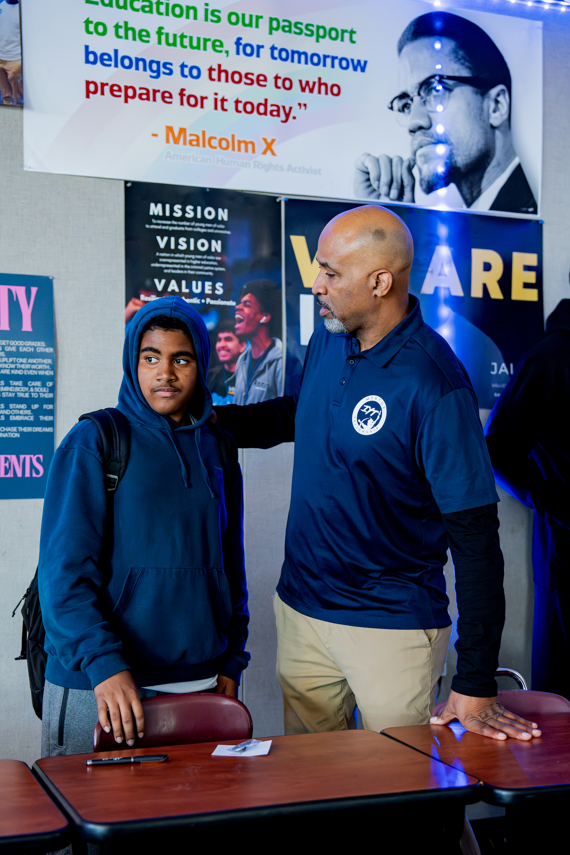 A young African American male student with a backpack, standing next to an adult man with a shaved head and a beard, wearing a blue button-up shirt and beige pants, holding a chair. They are in a room with various posters and a large motivational quote on the wall. The quote reads, "Education is our passport to the future, for tomorrow belongs to those who prepare for it today." - Malcolm X. The scene appears to be in an educational or community center setting.