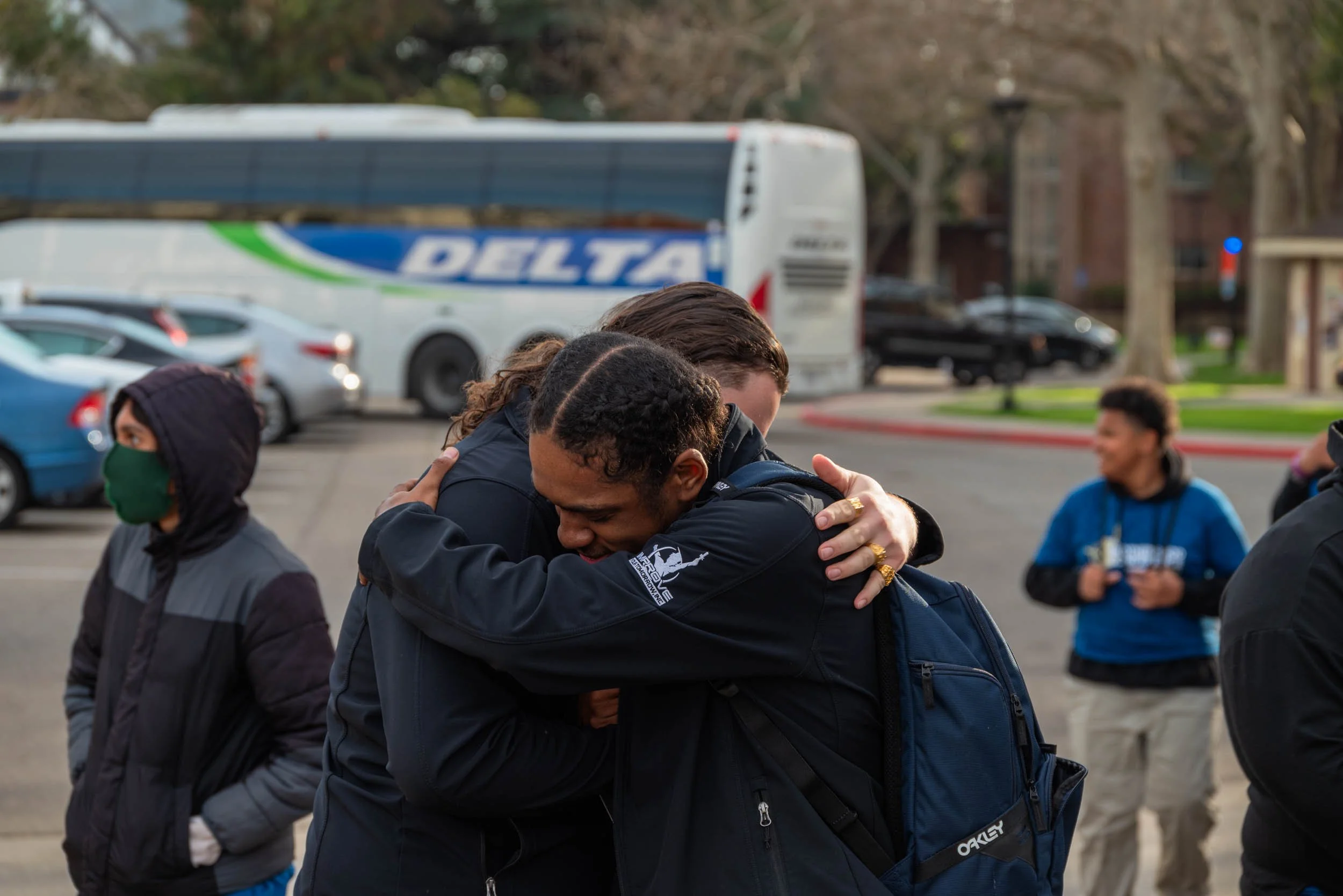 Group of people outdoors, hugging, with a Delta bus and parked cars in the background.