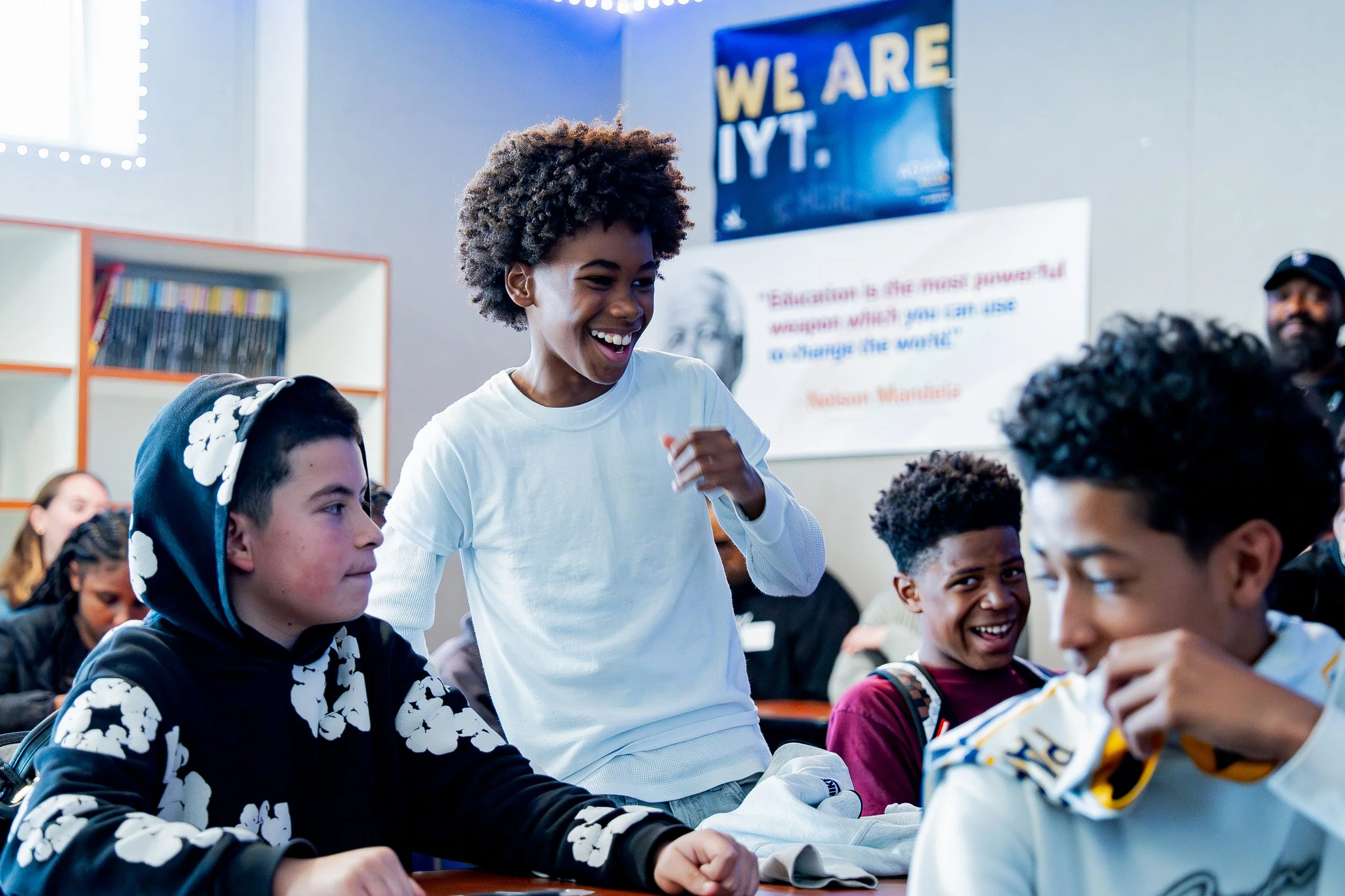 A group of students sitting in a classroom, with a boy standing and smiling, engaging with his classmates. The background includes educational posters and bookshelves.