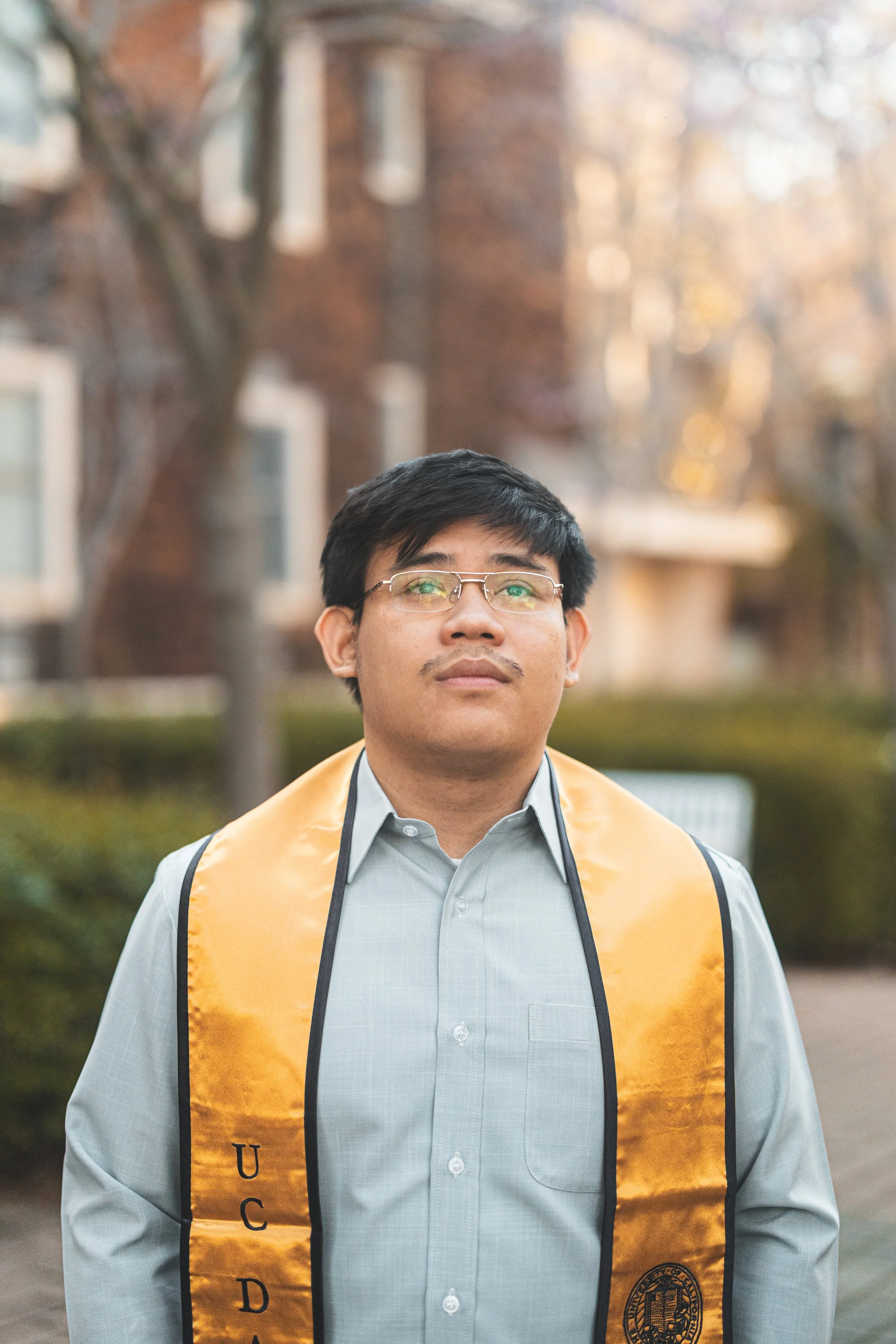 A young man wearing glasses and a gray dress shirt with a yellow graduation stole over his shoulders, standing outdoors in front of blurred trees and buildings, looking upward.