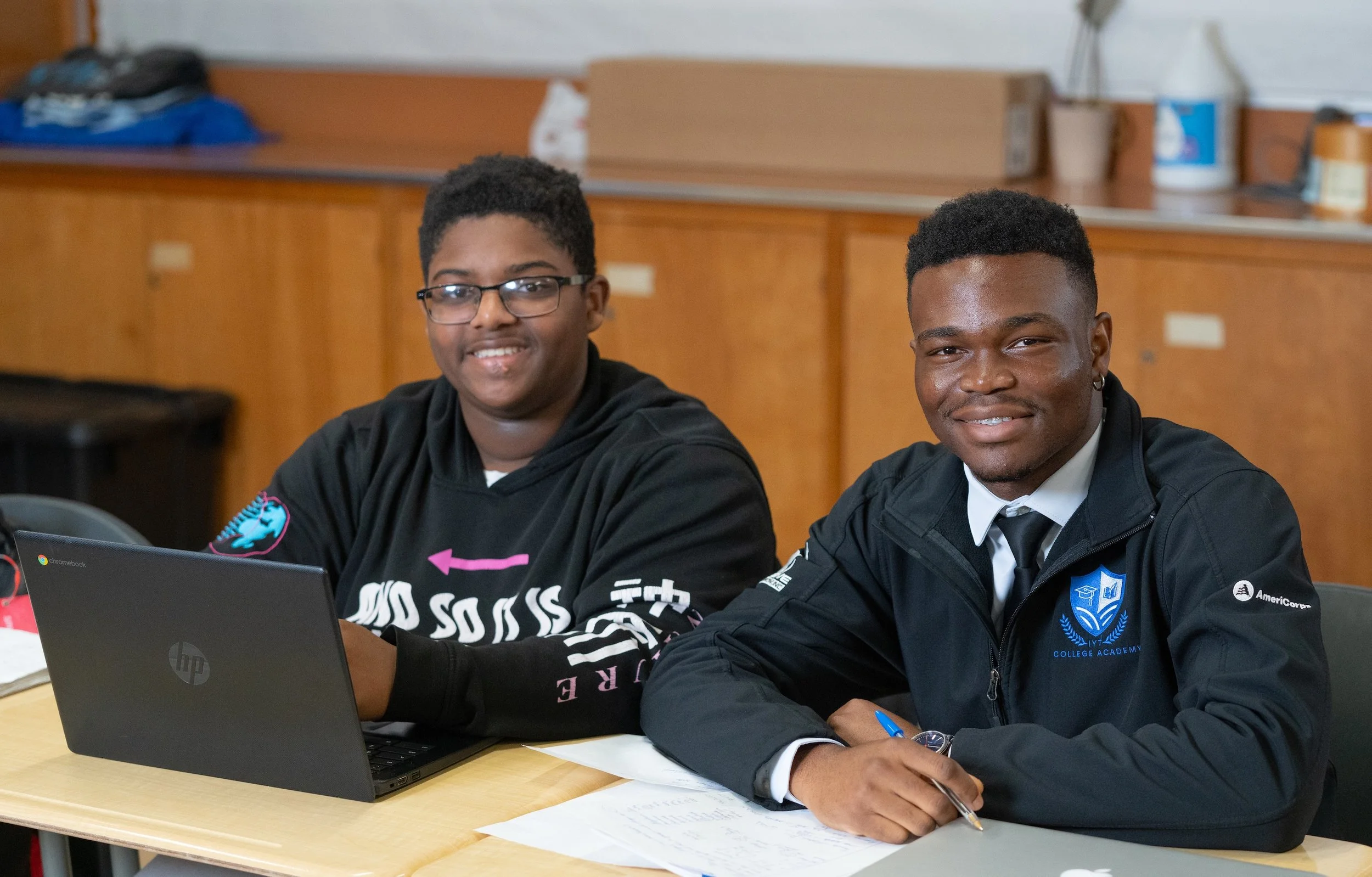 Four students sitting at a wooden table with laptops, focusing on their work in a classroom.
