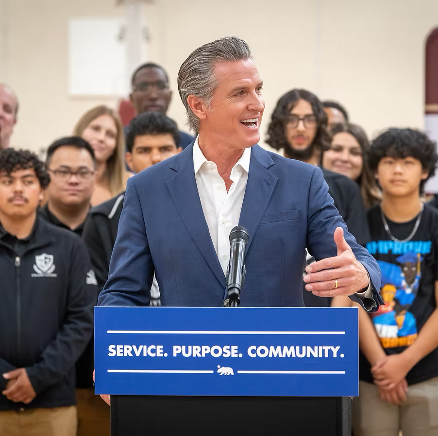 A man in a blue suit speaking at a podium with a group of young people standing behind him.