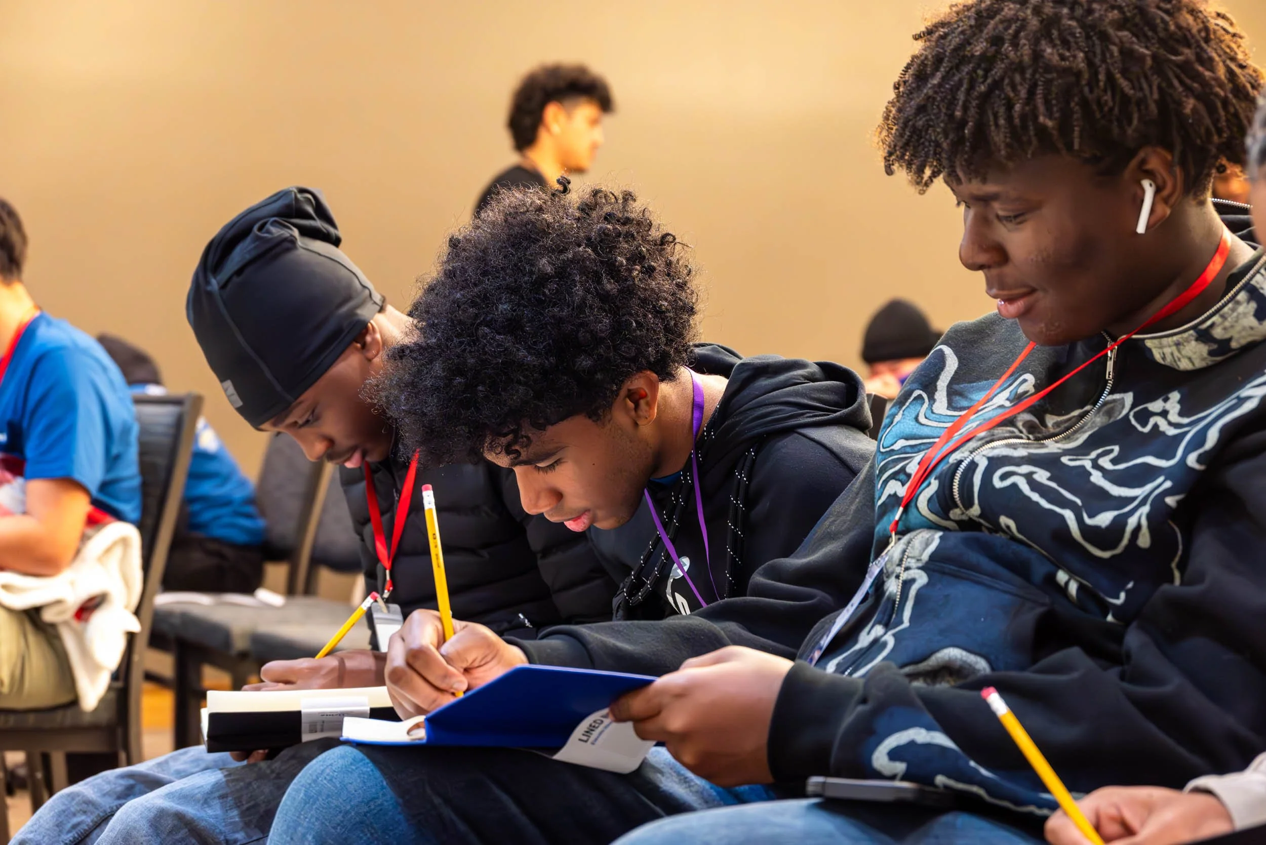 Group of young people sitting at a table, focused on writing or reading, in an indoor setting, some wearing headphones and lanyards.