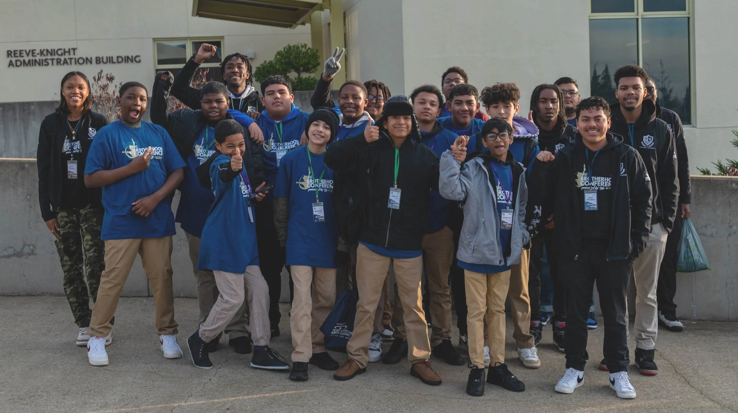 Group of diverse teenagers standing outdoors in front of the Reeve-Knight Administration Building, smiling and making gestures of excitement, wearing conference T-shirts and casual clothes.